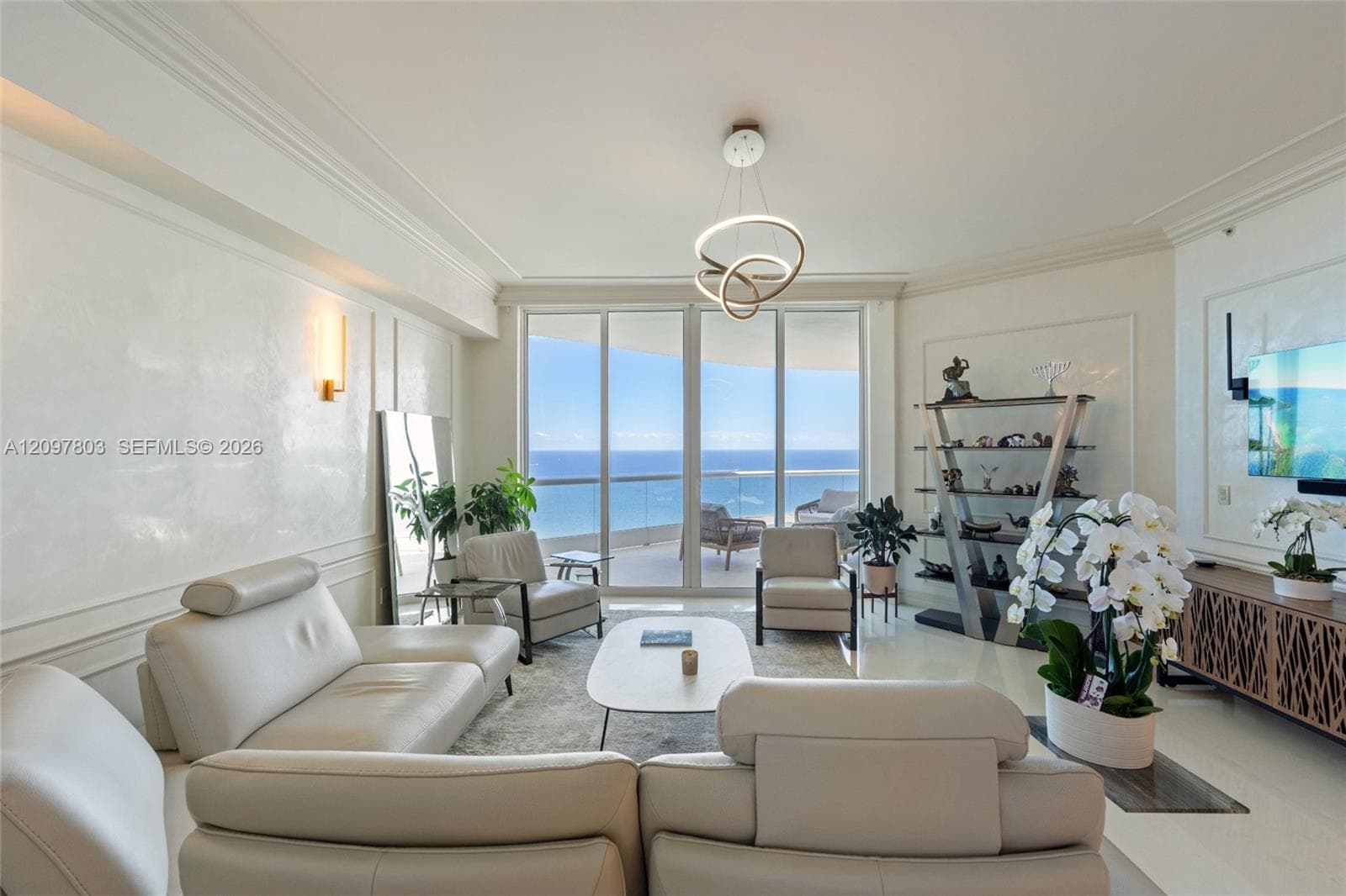 Living area featuring floor-to-ceiling glass sliding doors, polished stone flooring, intricate wall molding, modern sculptural lighting, and an attached balcony with glass railings