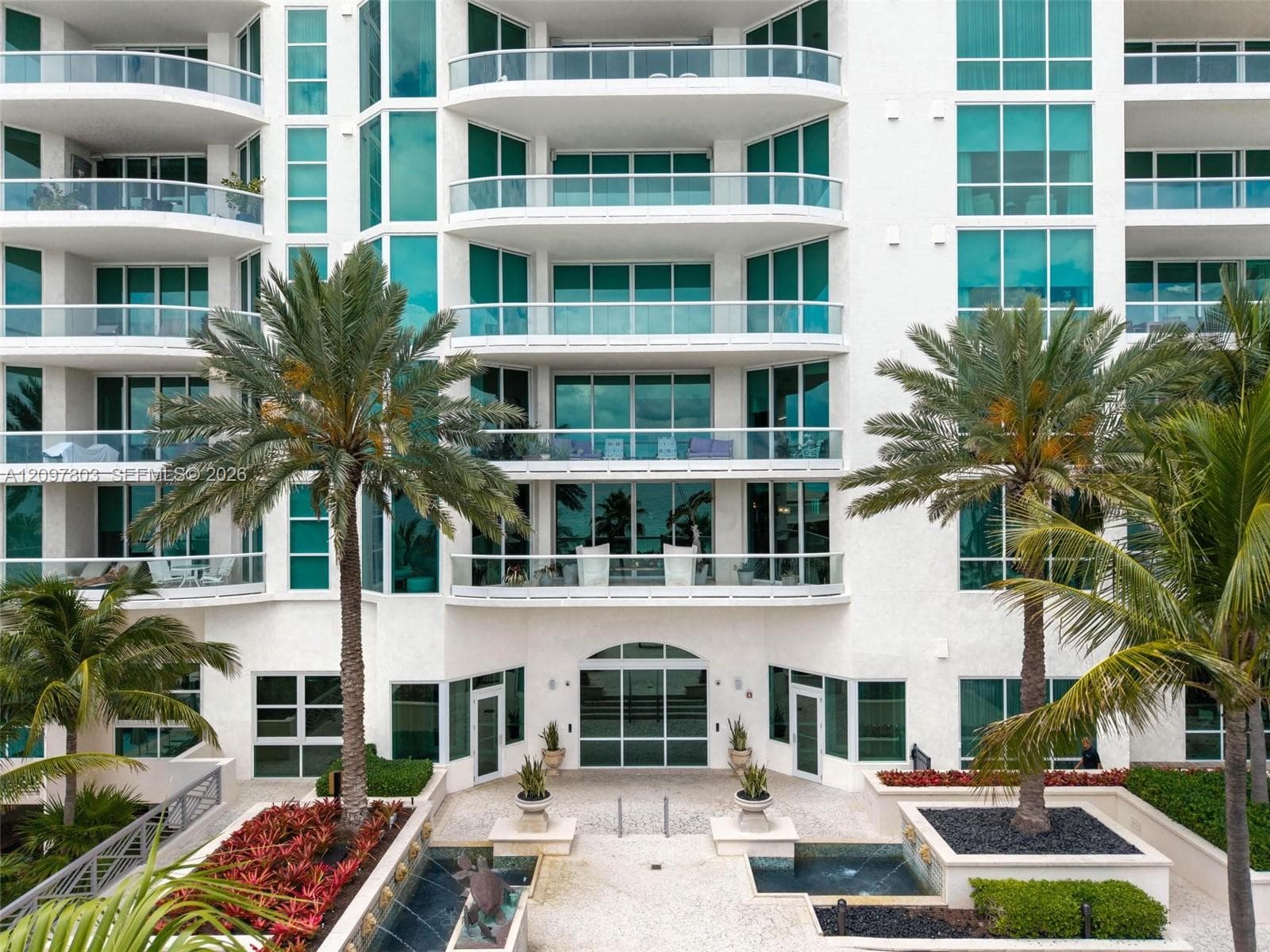 Multi-story high-rise exterior featuring a white stucco facade, full-height glass windows, private balconies with glass railings, and a landscaped courtyard with water features and mature palm trees