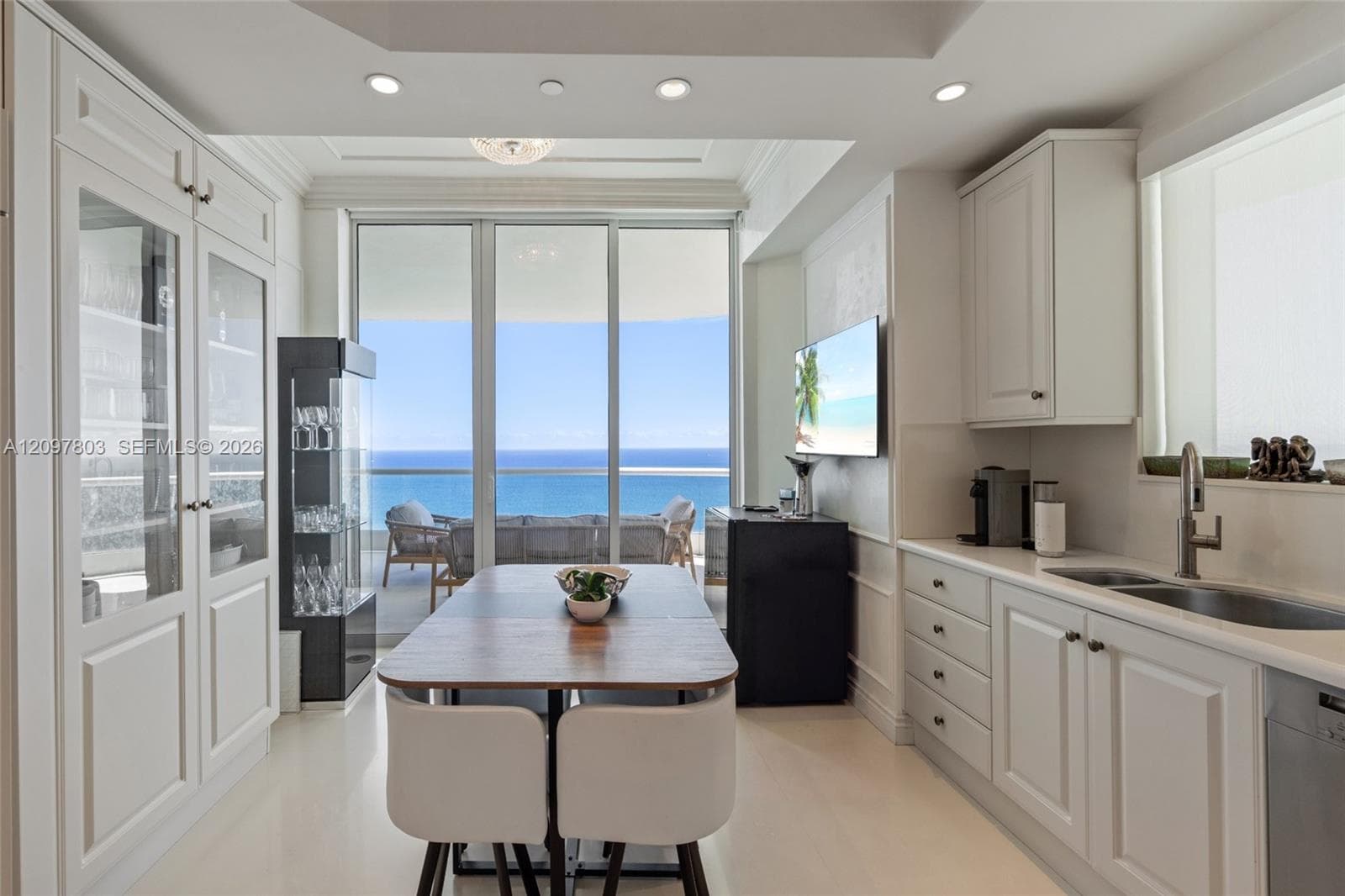 Custom cabinetry featuring glass-paneled display doors, recessed ceiling lighting, light-toned flooring, and floor-to-ceiling sliding glass doors opening to a balcony with ocean scenery