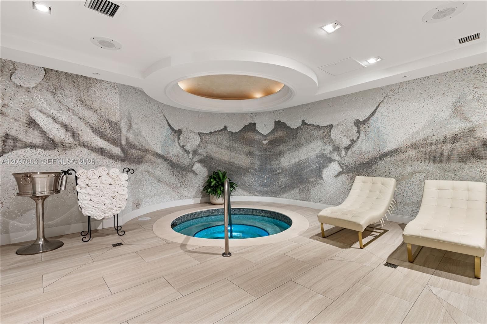 Custom-designed spa area featuring a circular tiled soaking pool, floor-to-ceiling mosaic accent wall, neutral large-format tile flooring, and a recessed ceiling cove with ambient lighting