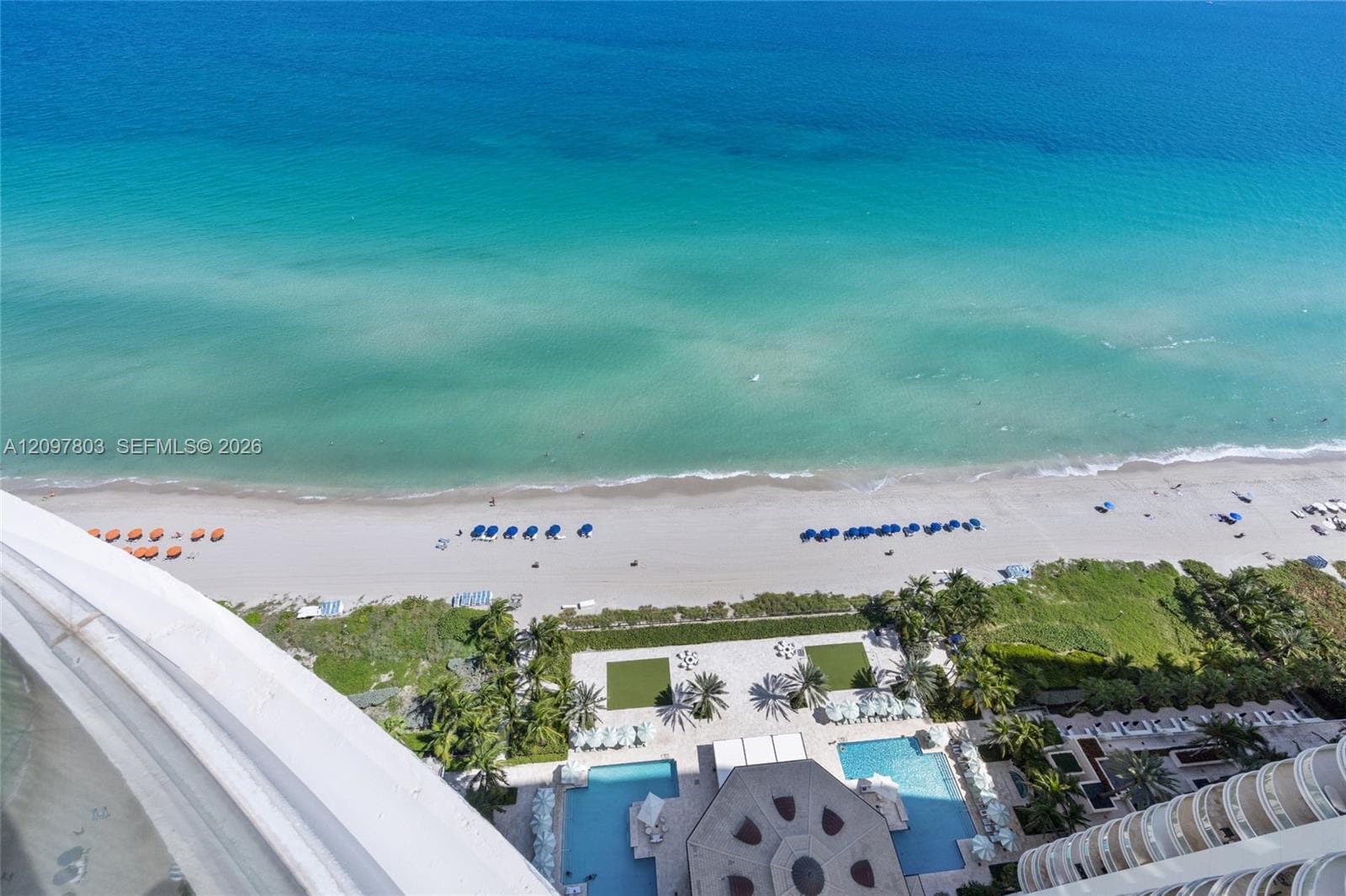 Direct oceanfront perspective featuring structural balcony railing, ground-level swimming pools, manicured green space, and sandy coastline