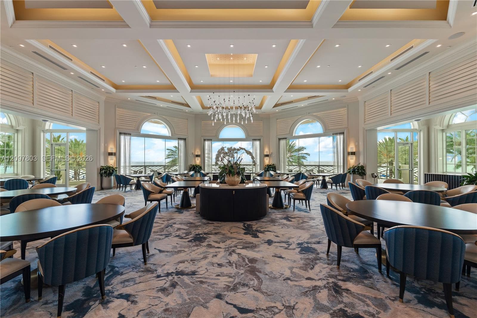 Expansive room featuring a coffered ceiling, intricate chandelier, recessed lighting, arched floor-to-ceiling windows, and integrated louvered shutters