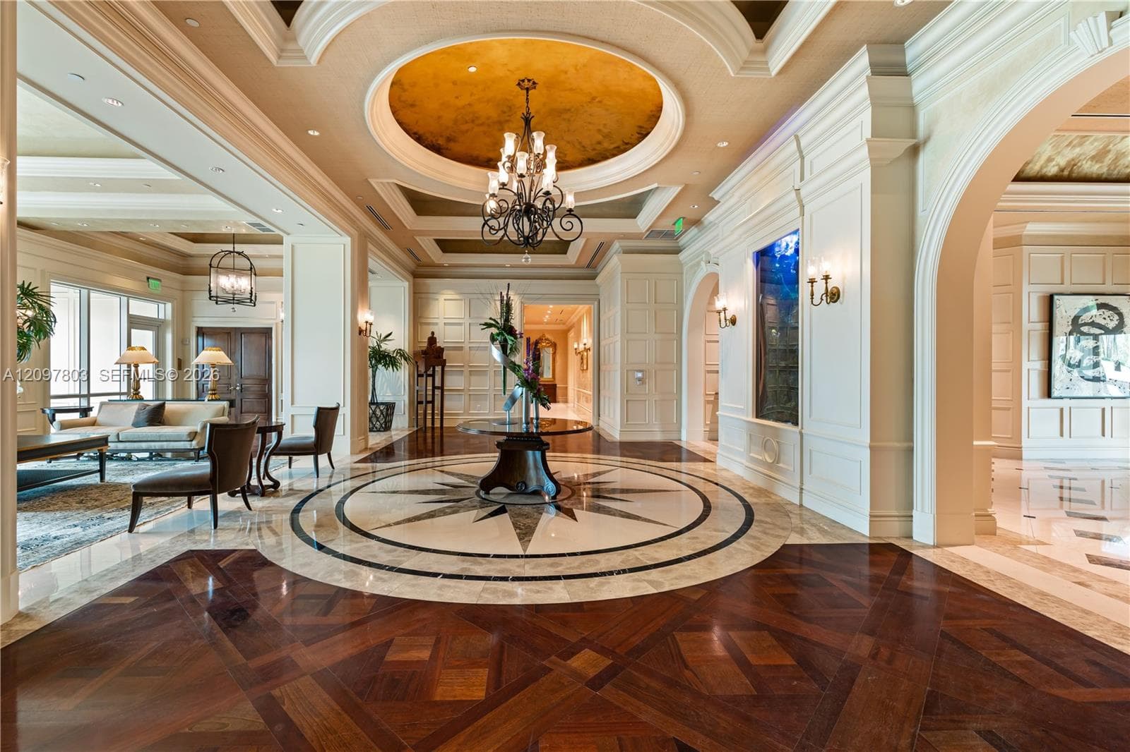 Grand foyer featuring a geometric wood-finish floor, a circular stone inlay medallion, coffered ceilings with metallic dome accents, integrated wall-mounted lighting, and paneled millwork