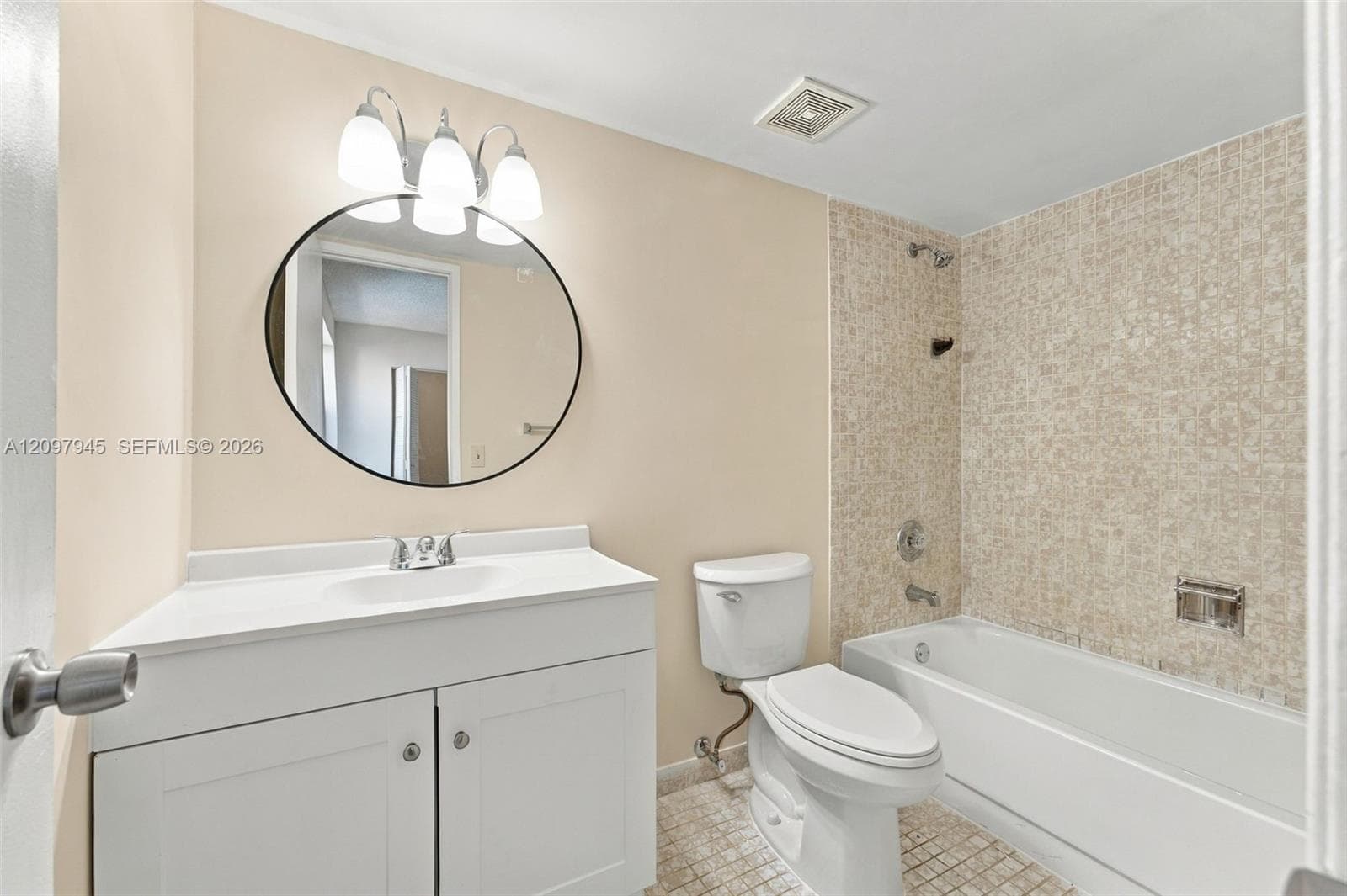 White vanity with integrated sink, circular wall-mounted mirror, three-light fixture, and bathtub with mosaic-tile surround