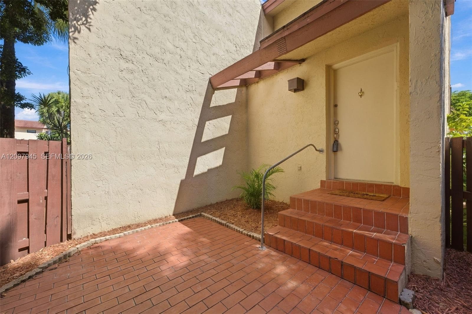 Exterior entryway featuring a red brick patio, matching brick steps, metal handrail, and stucco facade with a wood-accented awning