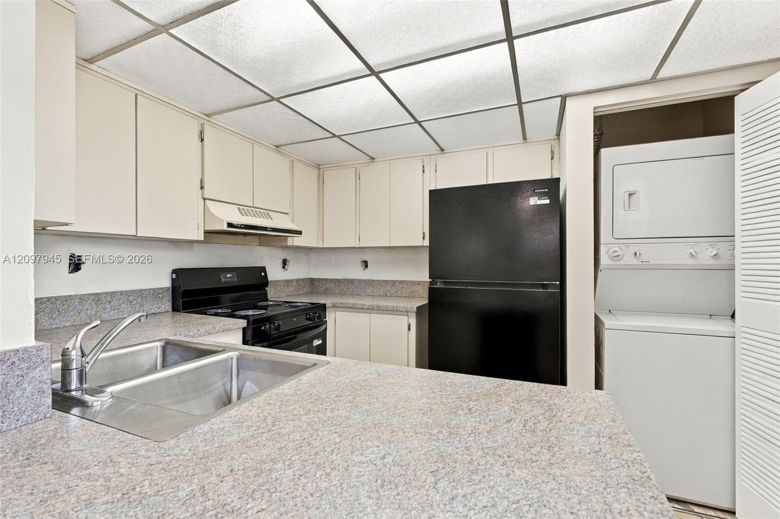 Kitchen featuring solid-surface countertops, dual-basin stainless steel sink, black range, black refrigerator, and built-in stacked laundry unit