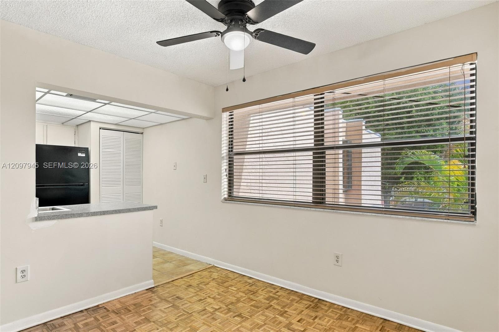 Parquet wood-finish flooring, ceiling fan with integrated lighting, large window with horizontal blinds, and a breakfast bar pass-through