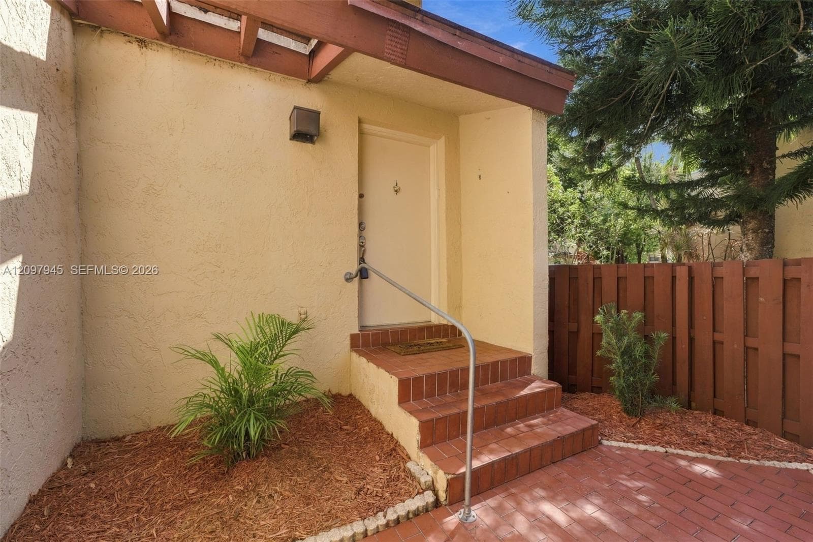 Exterior entryway featuring a stucco facade, covered porch with exposed timber beams, brick masonry steps, a metal handrail, and a private wooden privacy fence