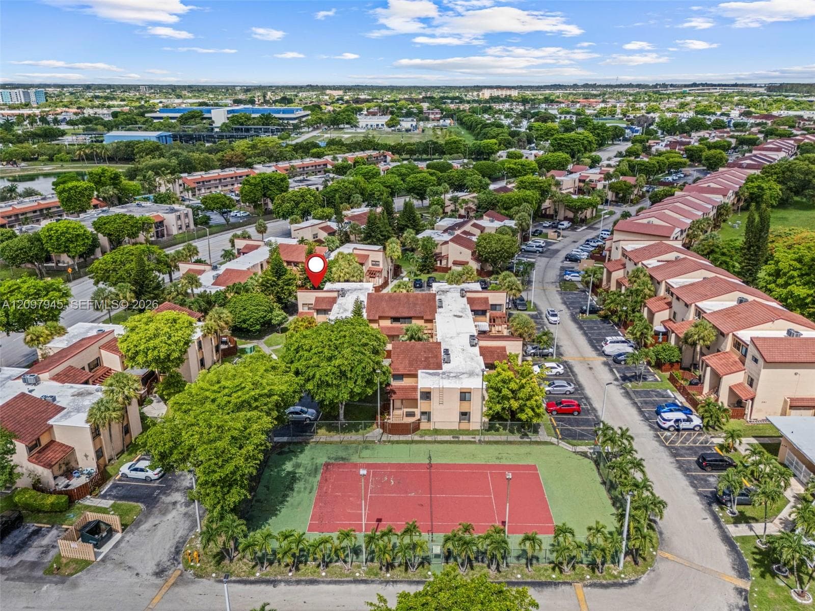 Aerial perspective of community grounds featuring a fenced tennis court, paved parking areas, and abundant mature landscaping