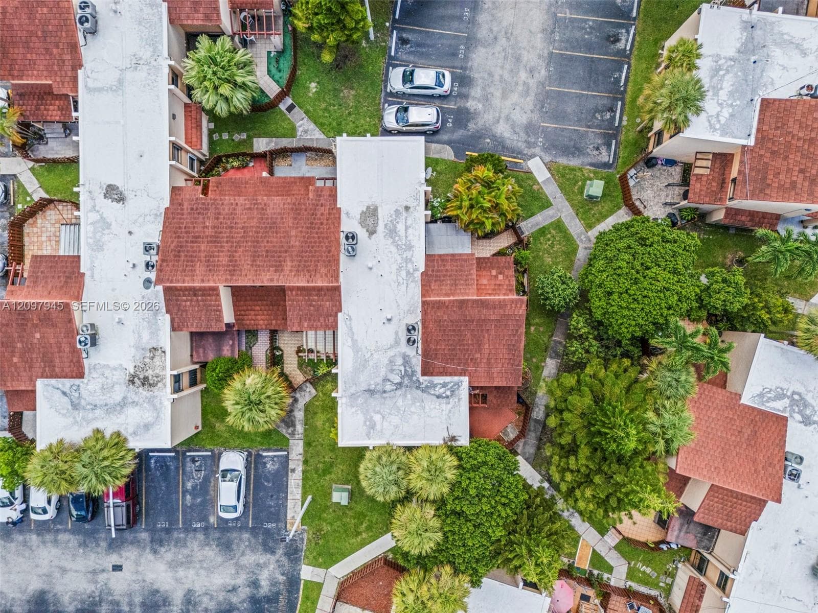Aerial perspective featuring multi-unit structures with shingle roofing, paved parking areas, concrete walkways, and mature palm trees