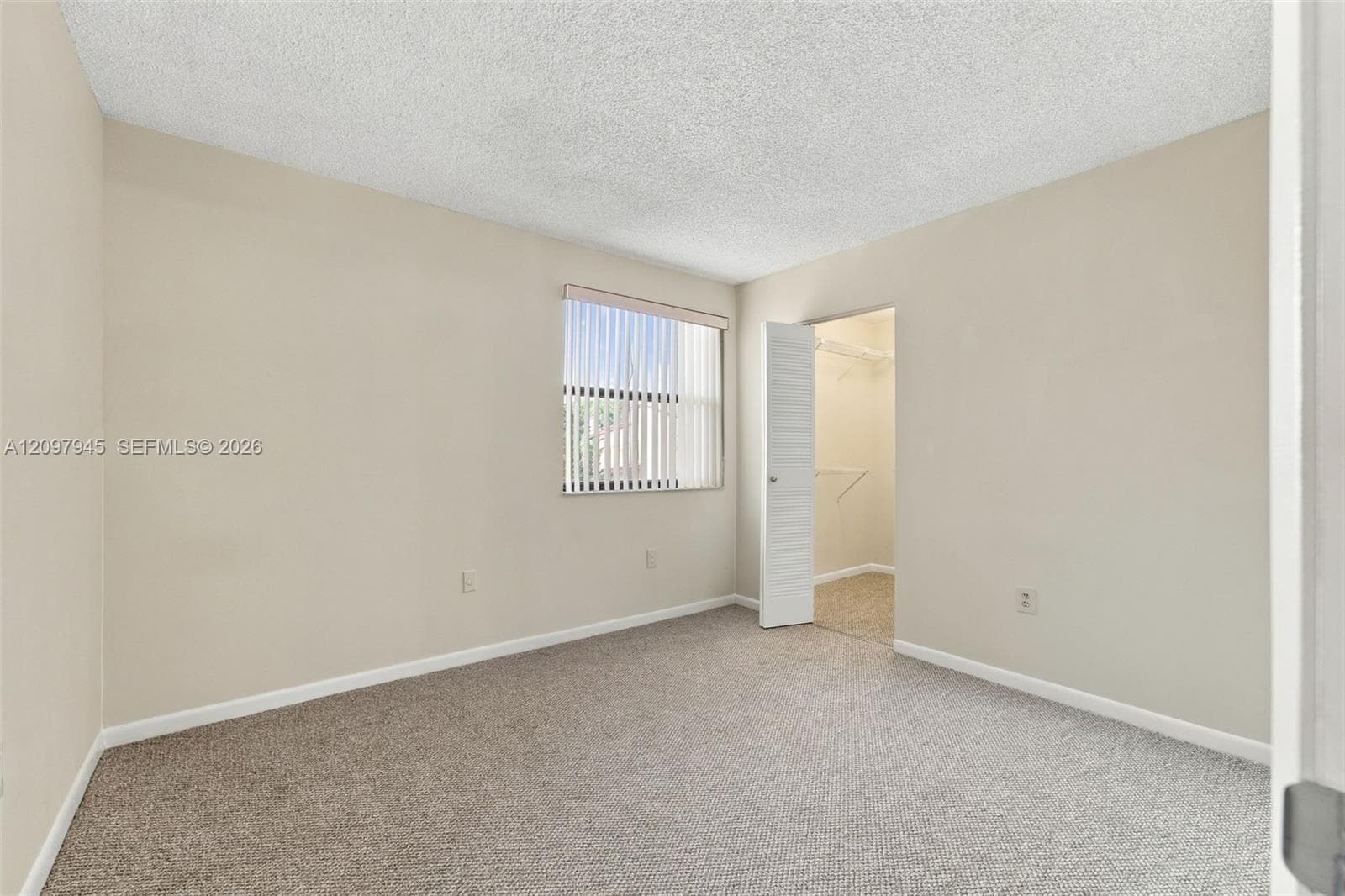 Neutral-toned interior featuring wall-to-wall carpeting, textured ceiling, single window with vertical blinds, and a walk-in closet with built-in shelving