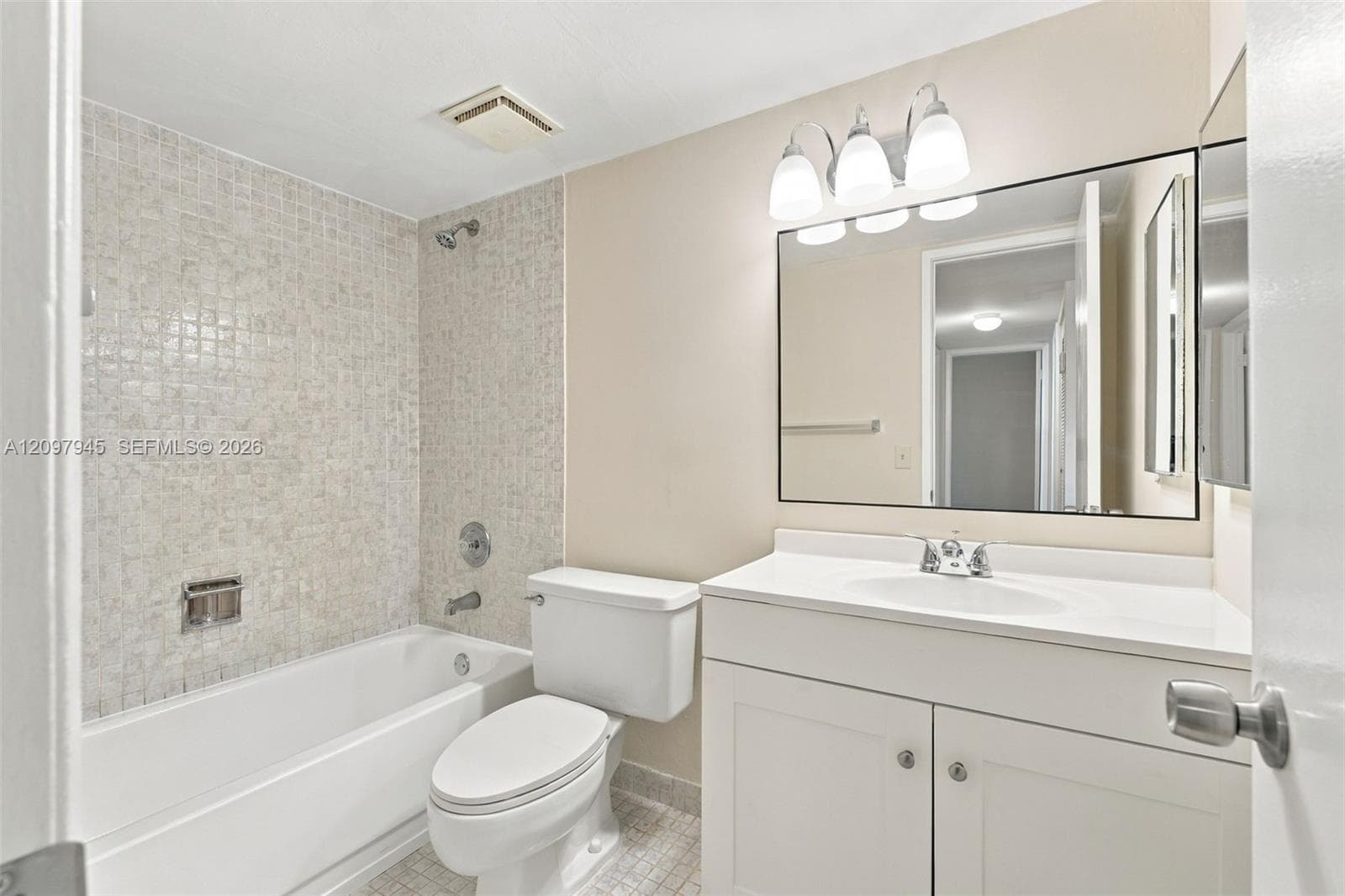 Bathroom featuring a white vanity with a composite countertop and integrated sink, a large wall-mounted mirror, a three-light fixture, and a shower-tub combination finished with light-toned mosaic tile surround