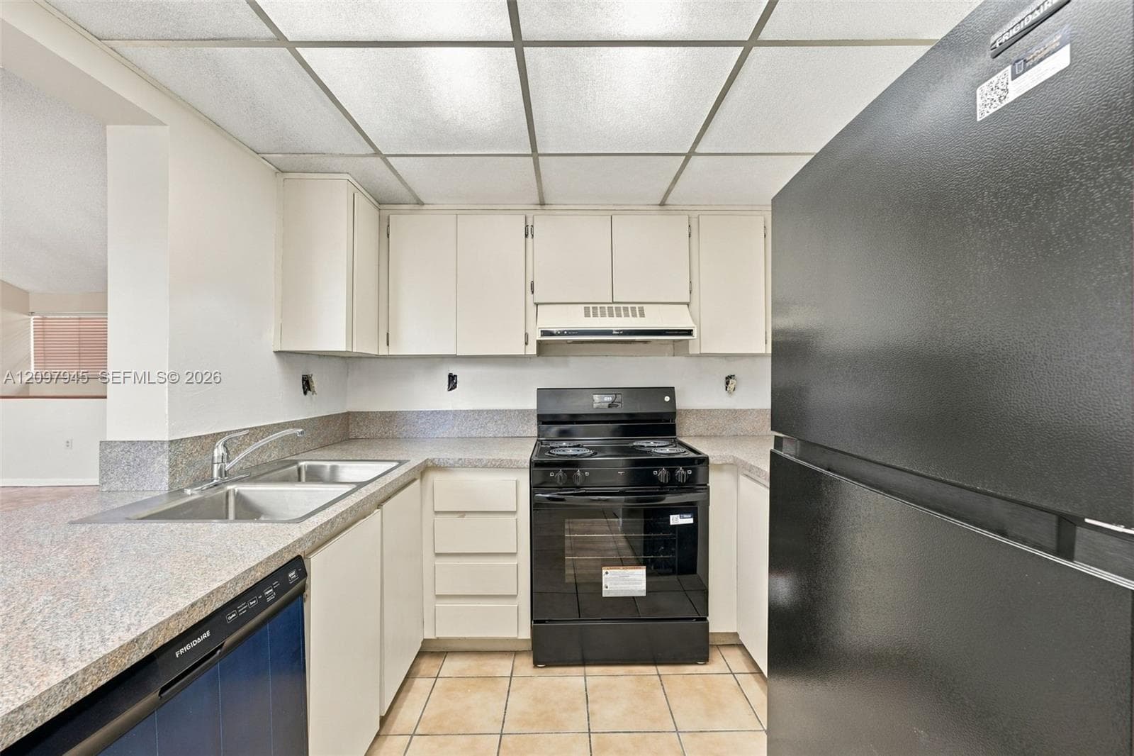 Kitchen featuring white cabinetry, solid-surface countertops, dual-basin stainless steel sink, tile flooring, and black appliances