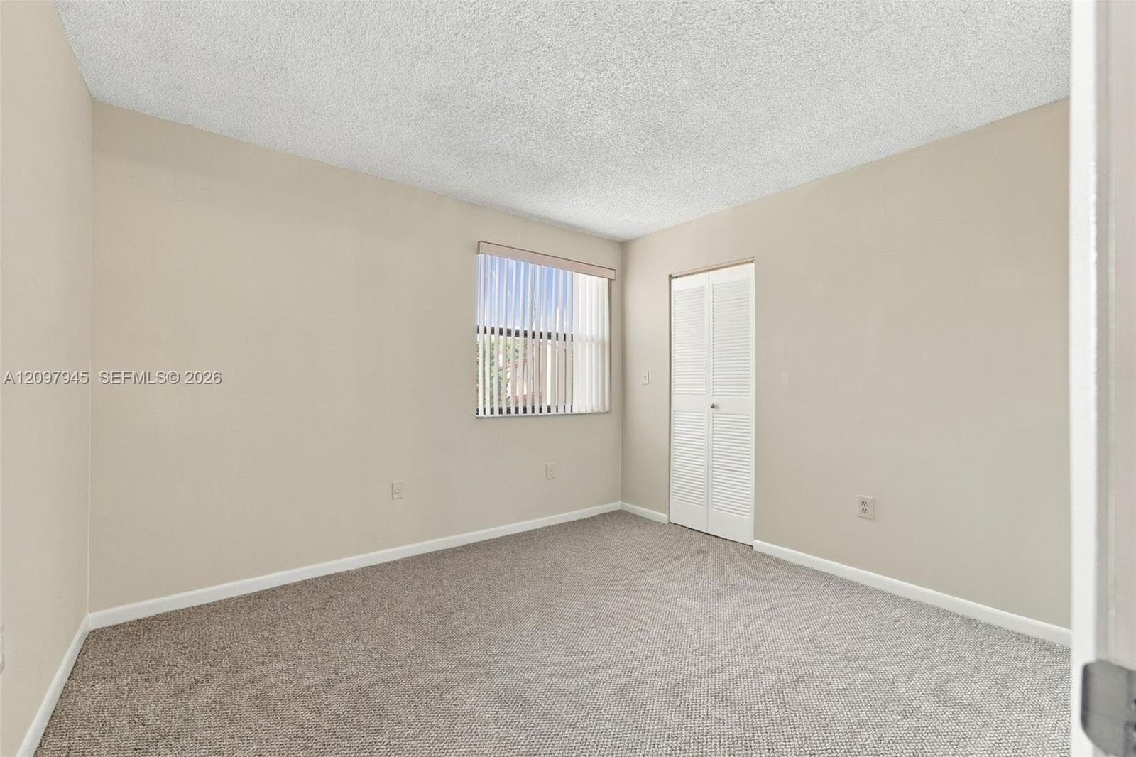Room interior featuring neutral-toned walls, textured ceiling, carpeted flooring, a single-pane window with vertical blinds, and a built-in closet with double bifold doors