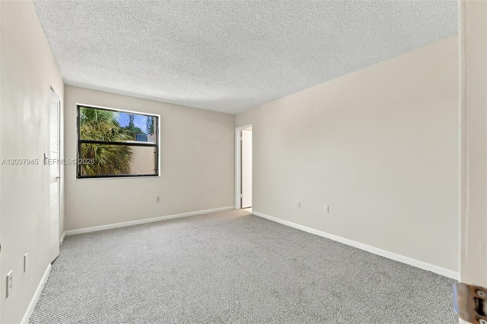 Neutral interior room featuring neutral wall paint, carpeted flooring, textured ceiling, and a single-pane window with a louvered closet door
