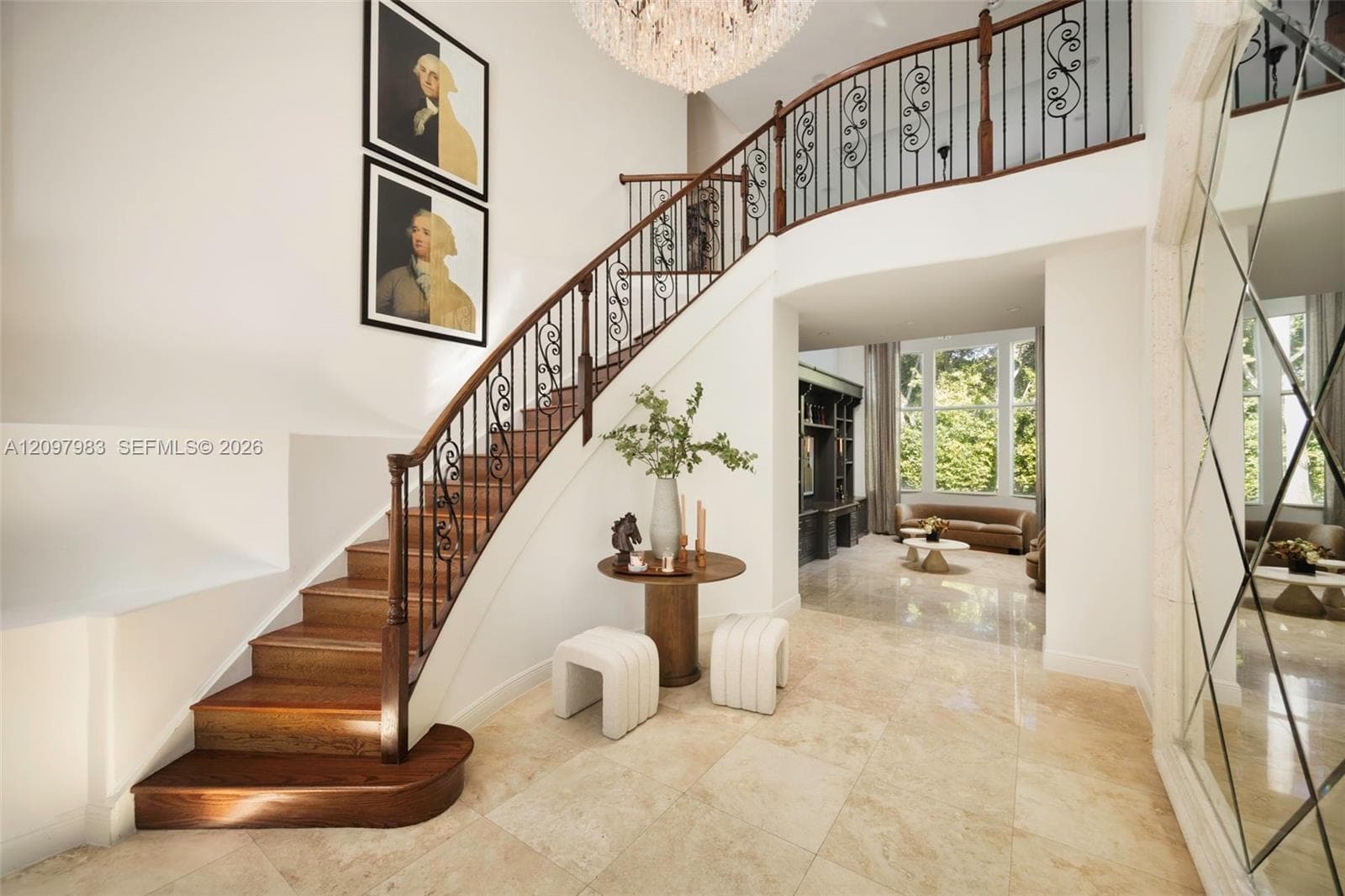 Grand foyer featuring natural stone flooring, a sweeping staircase with custom metal balusters and wood handrails, a multi-tiered crystal chandelier, and a floor-to-ceiling diamond-patterned mirror