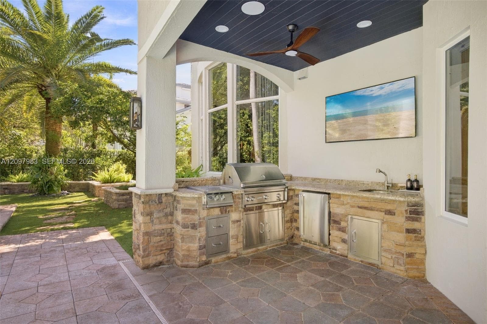 Covered patio featuring an outdoor kitchen with stainless steel appliances, stacked stone base, stone countertop, integrated sink, and recessed ceiling lighting