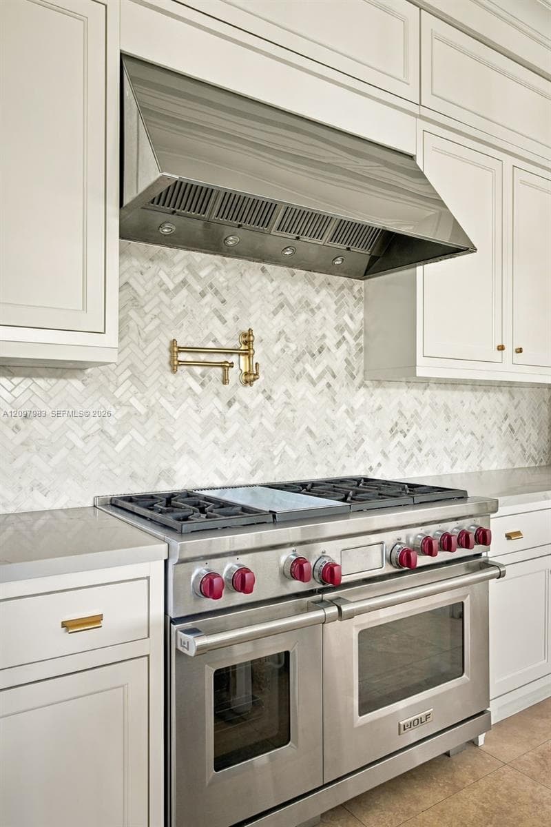 Professional-grade stainless steel range with gas cooktop, herringbone-patterned stone backsplash, gold-tone wall-mounted pot filler, custom shaker-style cabinetry, and stainless steel range hood