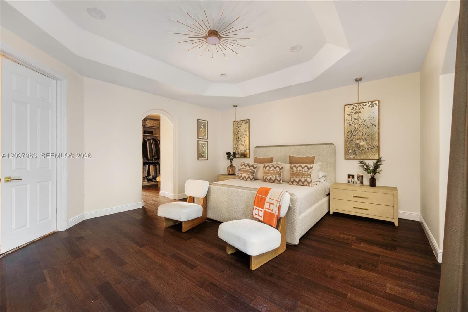 Tray ceiling with central sunburst-style fixture, wood-finish flooring, and arched entryway to a walk-in closet