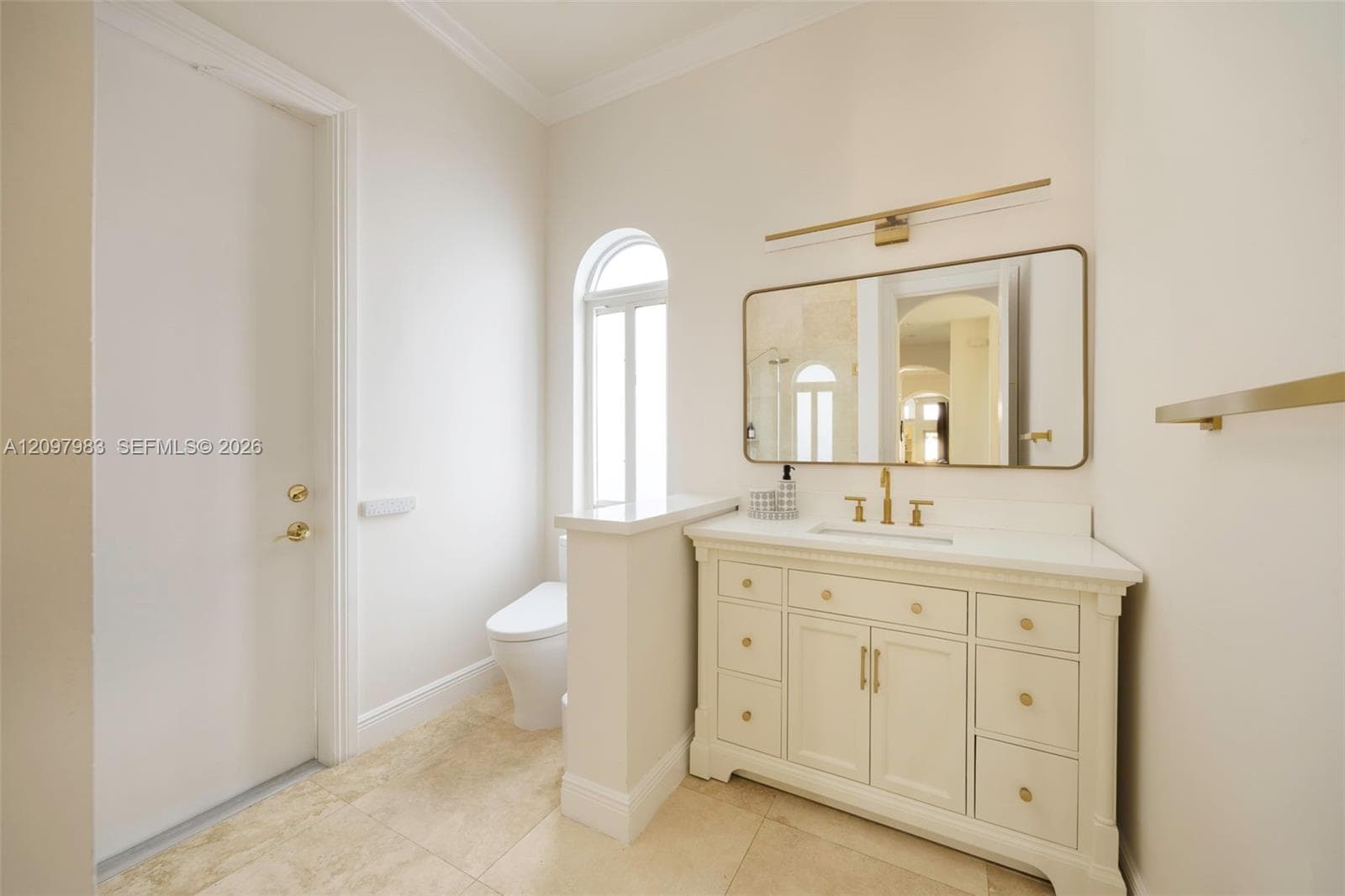 Bathroom featuring a white vanity with gold-tone hardware, stone-finish tile flooring, an arched window, and integrated vanity lighting
