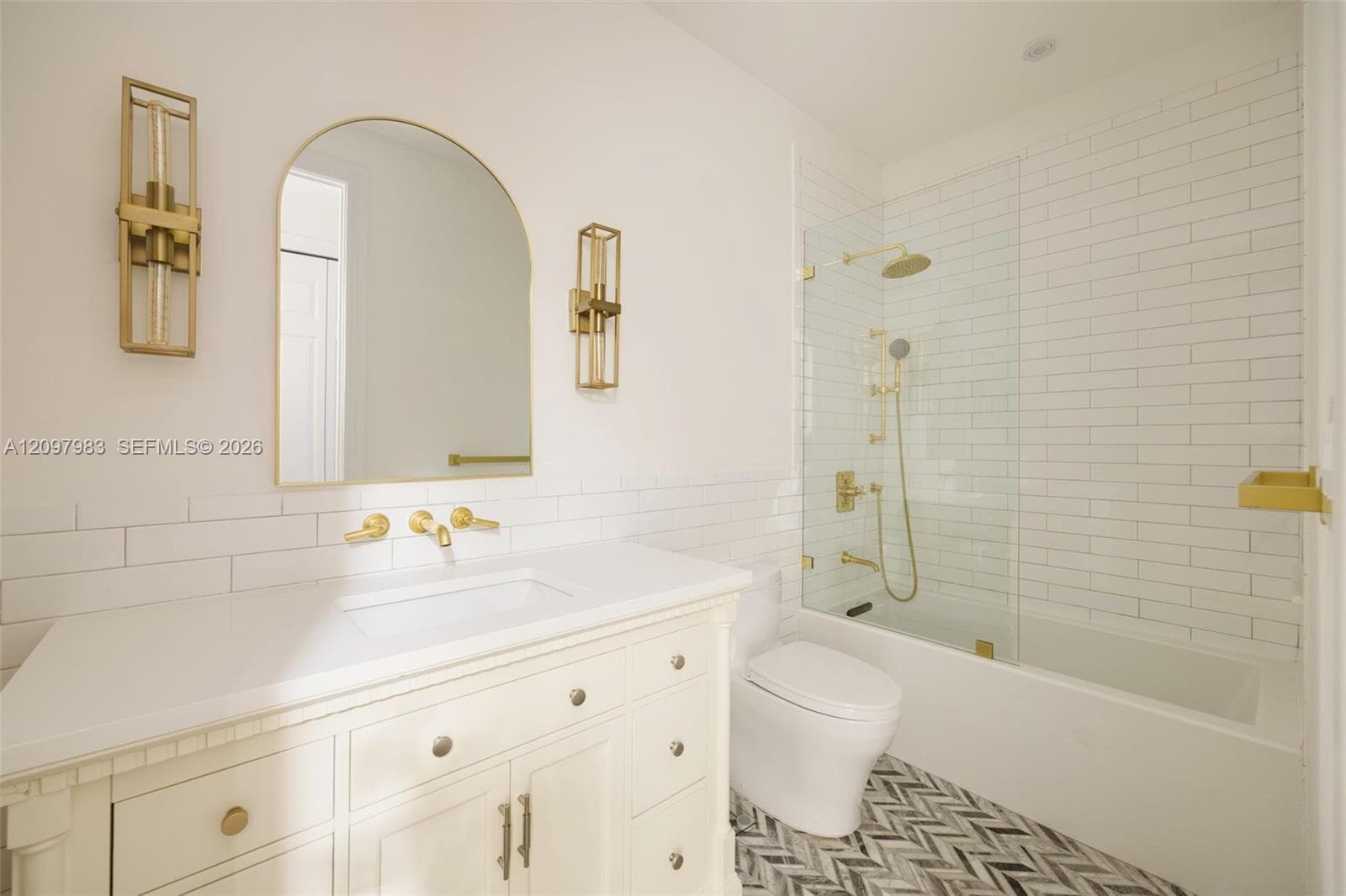 Bathroom featuring a white vanity with countertop, arched wall mirror, metallic gold-tone fixtures, glass-enclosed shower with subway tile surround, and patterned floor tiling