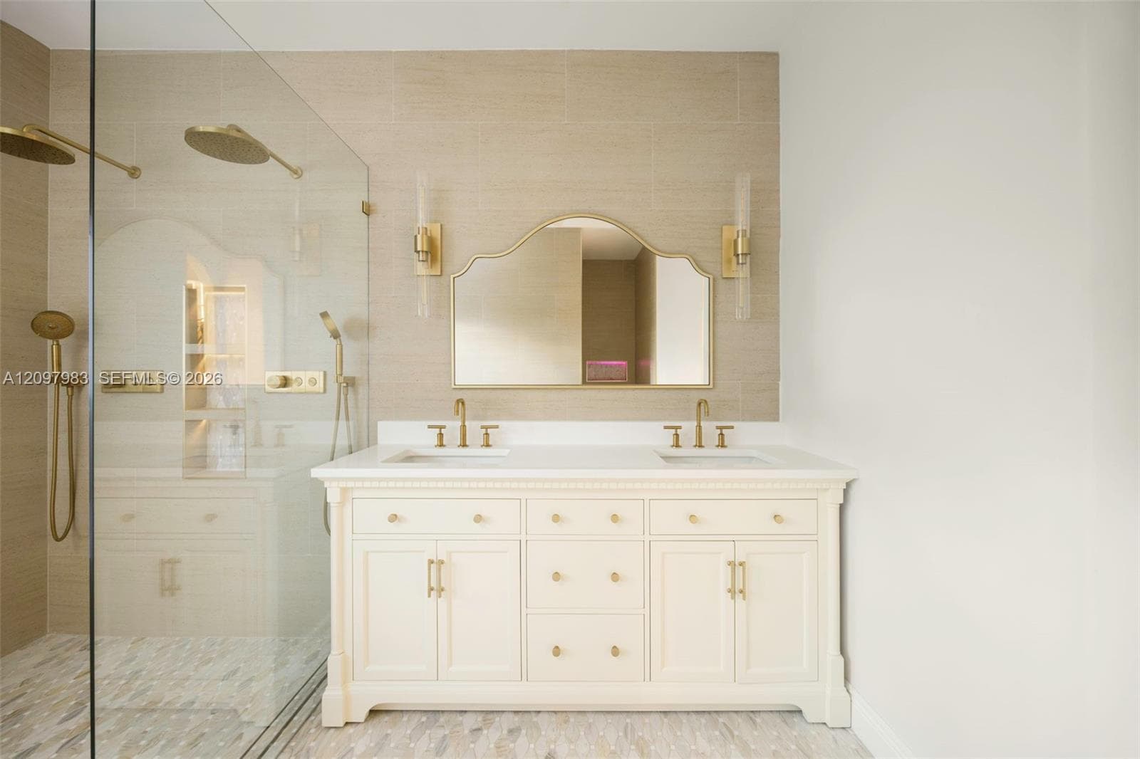 Double vanity with a white countertop and gold-toned hardware, arched wall mirror, wall-mounted brass light fixtures, and a glass-enclosed shower featuring dual rainfall showerheads and horizontal tile wall surrounds