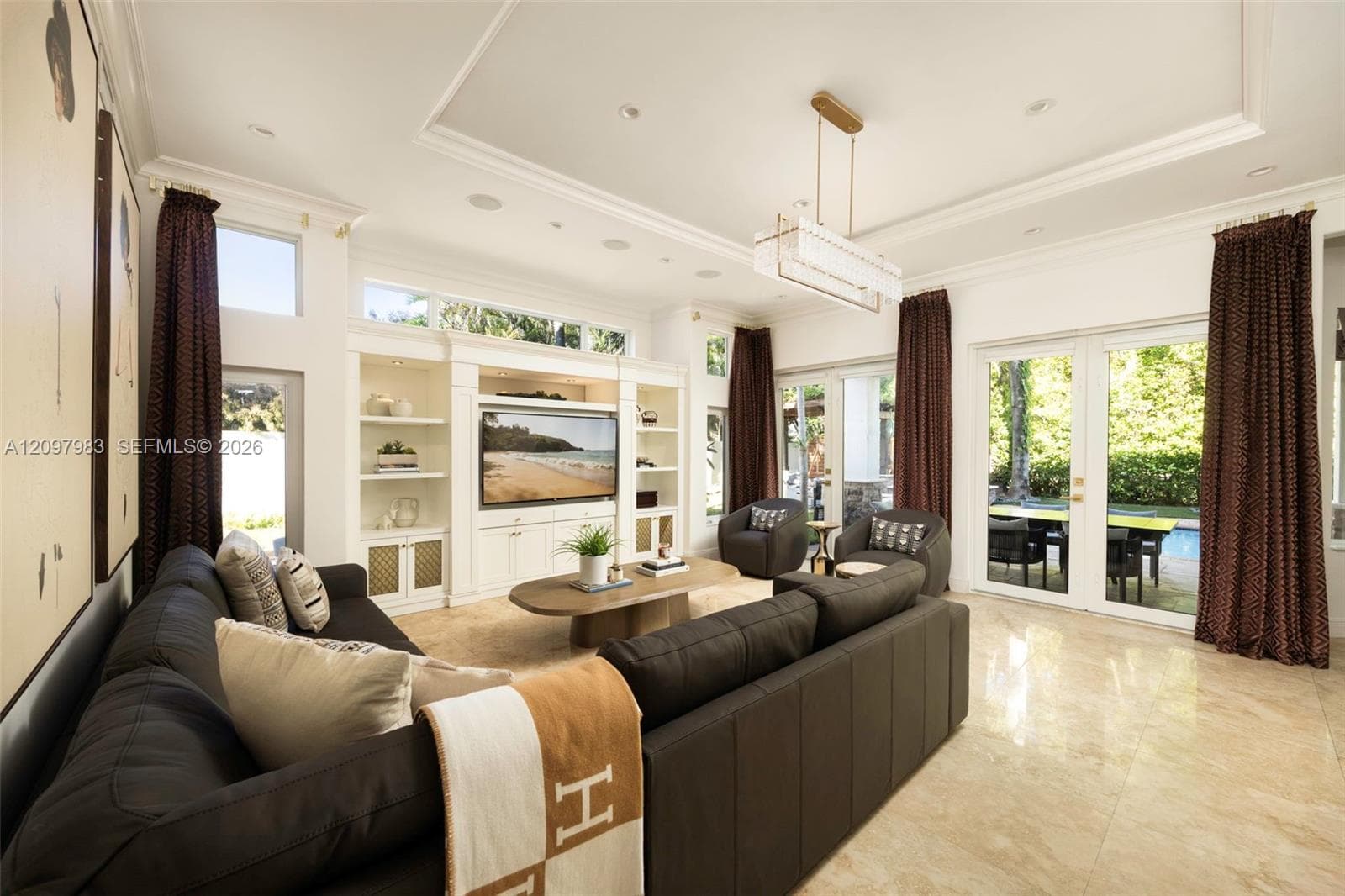 Living area featuring a custom built-in entertainment center, tray ceiling with recessed lighting, stone-finish tile flooring, and oversized glass-paneled double doors