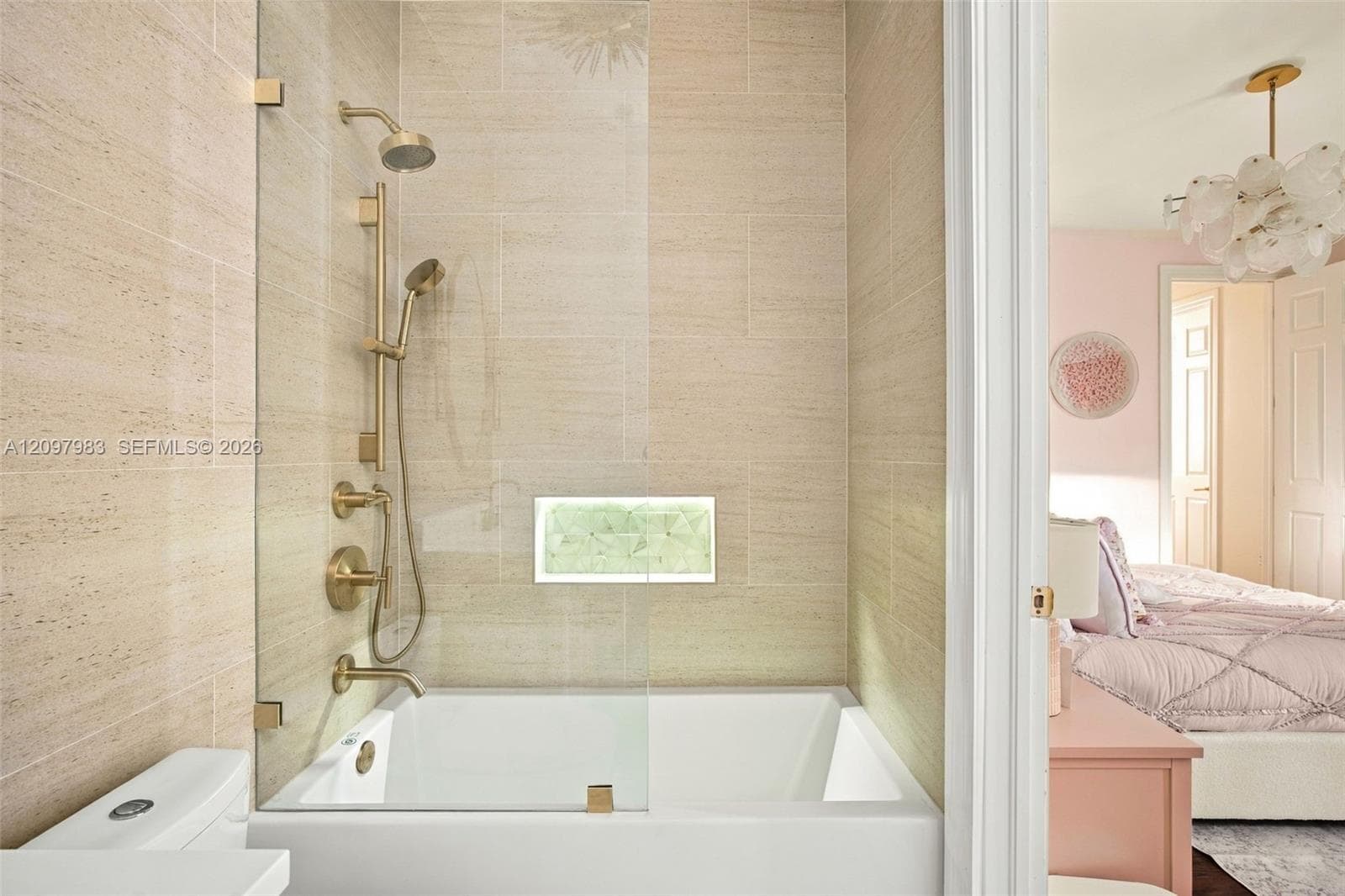 Bathtub with a glass panel, brass-finish rainfall showerhead and handheld sprayer, stone-tile wall surround, and a recessed tiled niche