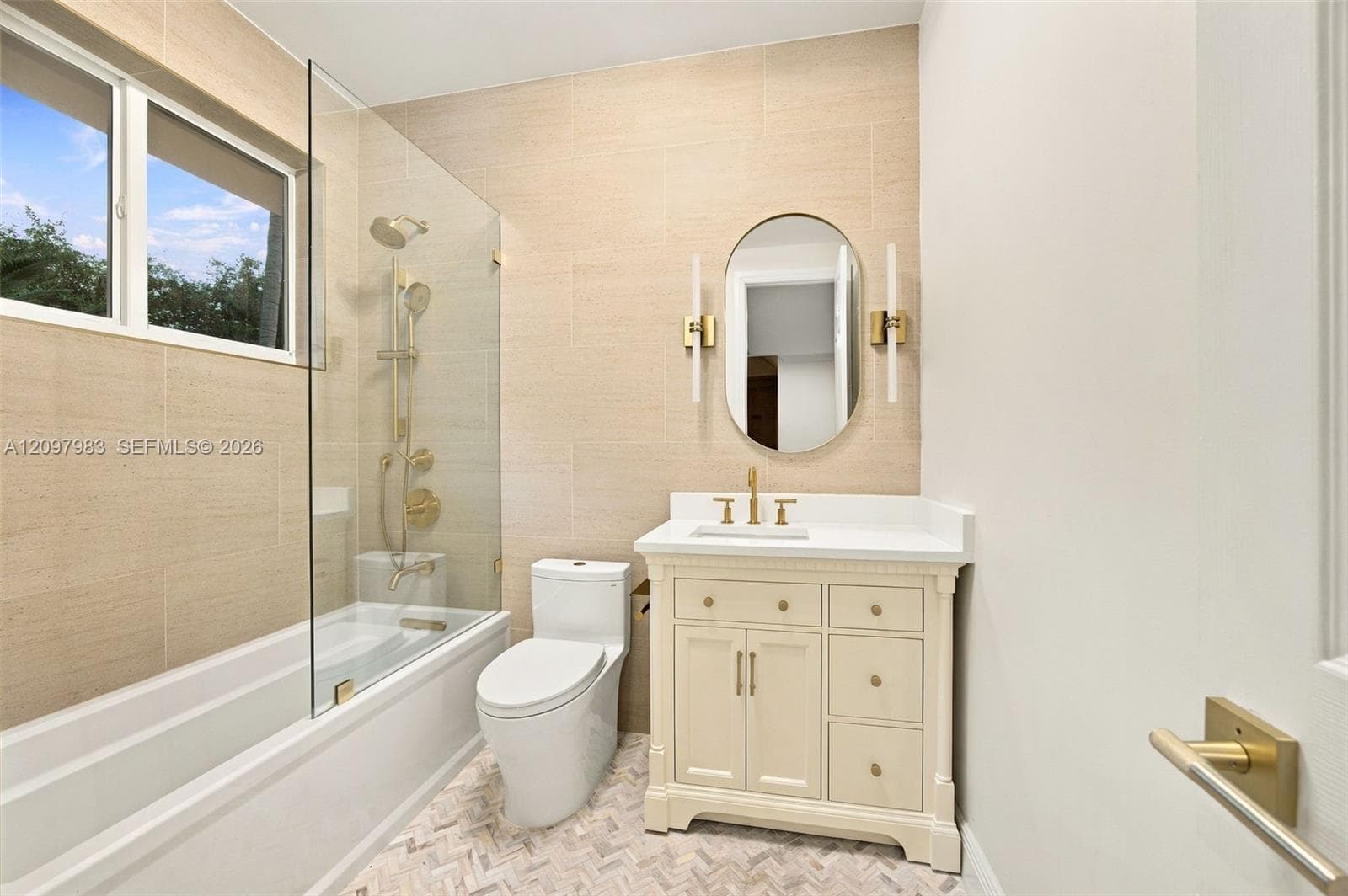 Bathroom featuring a cream-colored vanity with a white countertop, oval mirror with brass-finish wall sconces, glass-enclosed shower and tub combination with brass-finish fixtures, and neutral textured wall tile