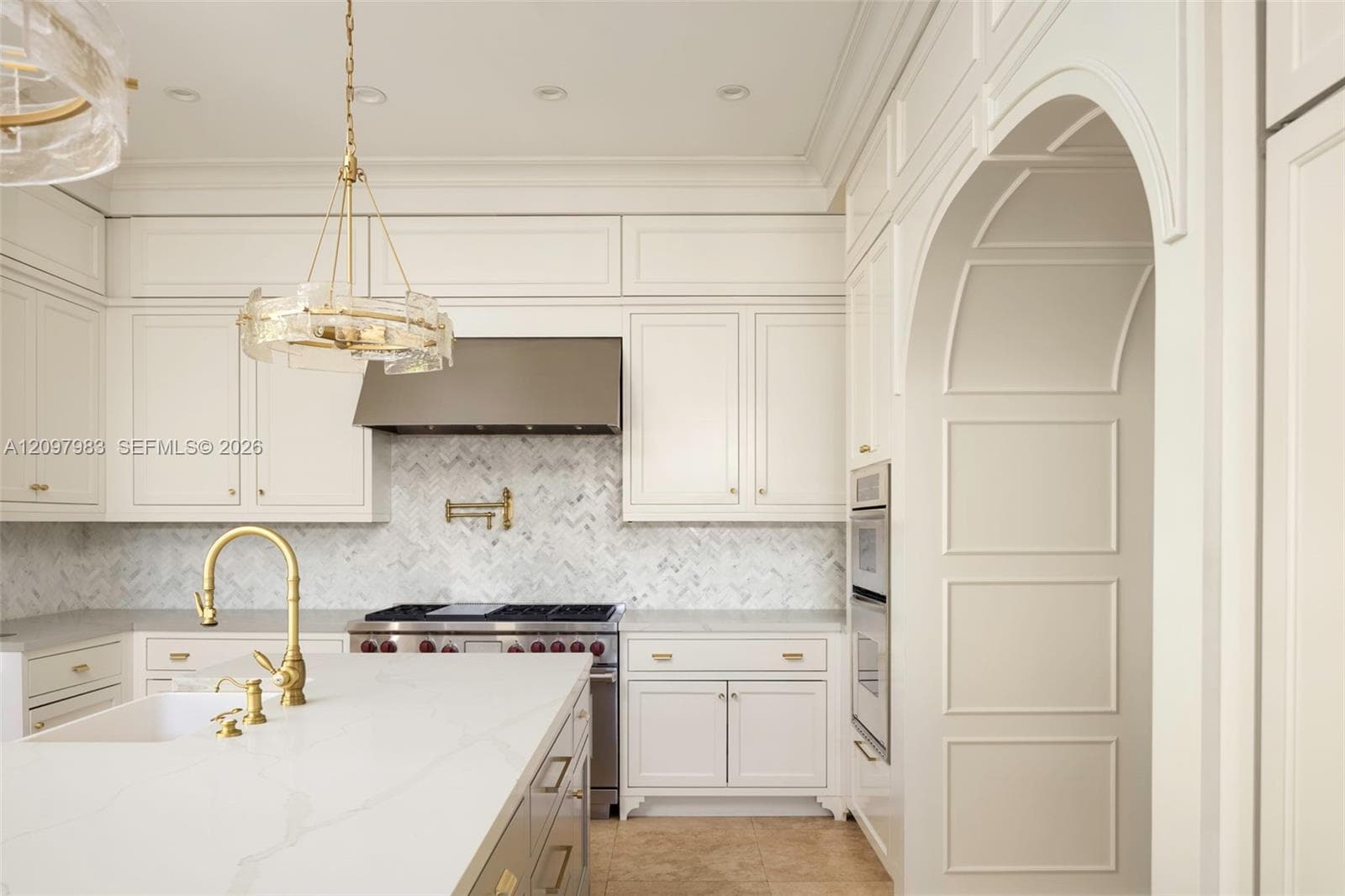Kitchen featuring custom cabinetry, herringbone-pattern stone tile backsplash, brass-finish fixtures, quartz countertops, and a paneled arched entryway