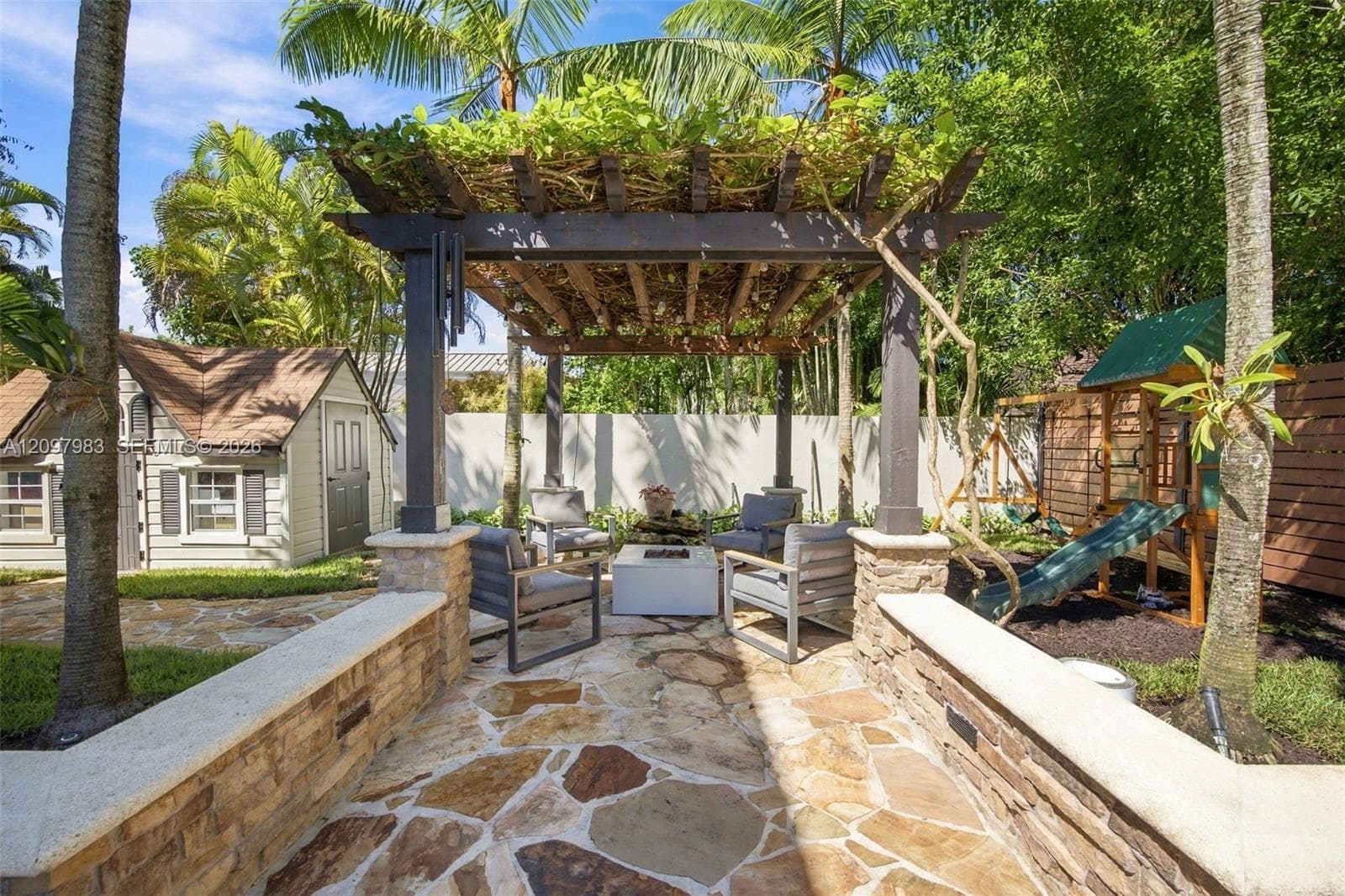 Outdoor hardscape featuring a stone-paver patio, a wooden pergola with integrated lighting, and stone-clad retaining walls with capstones