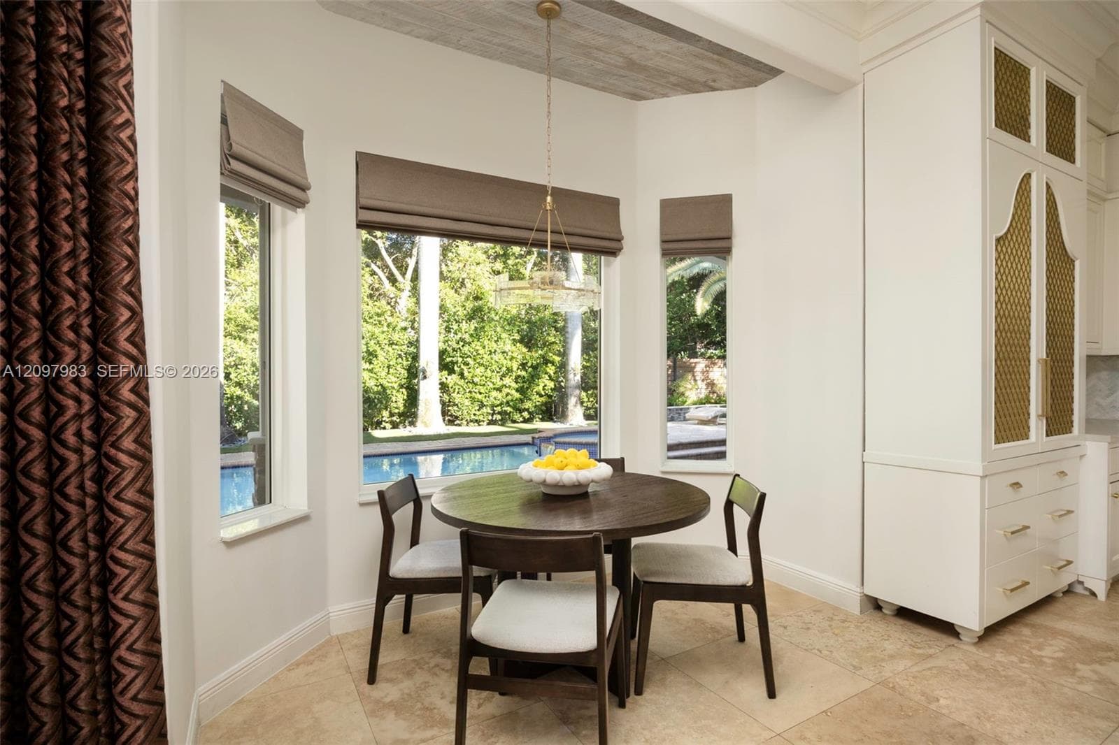 Sun-drenched nook featuring tile flooring, custom ceiling beam detailing, floor-to-ceiling built-in cabinetry with decorative metal panel inserts, and large-scale windows with exterior pool exposure