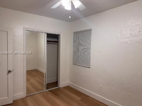 Room featuring wood-finish flooring, sliding mirrored closet doors, a multi-blade ceiling fan with light fixture, and horizontal window blinds