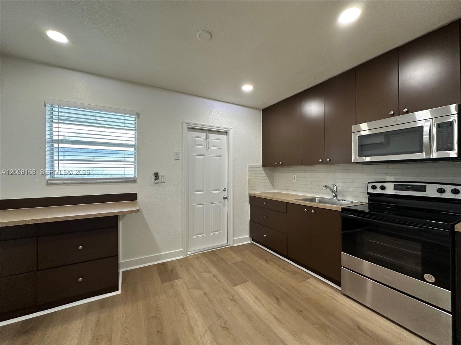 Kitchen featuring dark wood-finish cabinetry, stainless steel appliances, neutral stone-finish countertops, subway tile backsplash, and wood-finish flooring