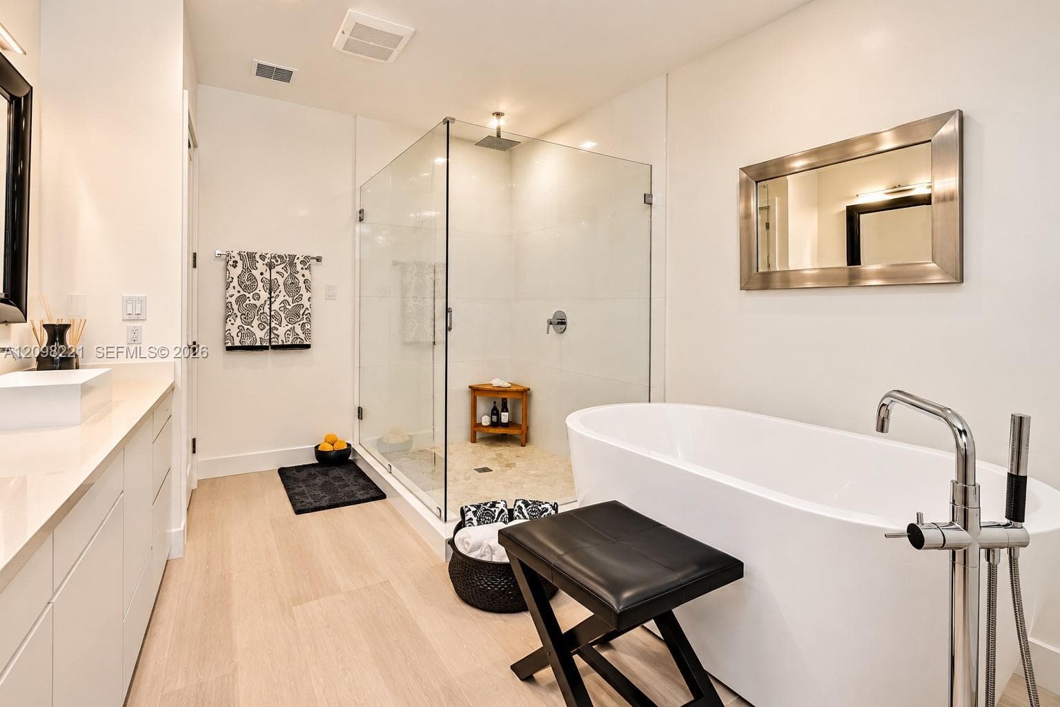 Freestanding soaking tub with floor-mounted chrome faucet, glass-enclosed shower with stone flooring, wall-mounted mirror, and smooth-finish cabinetry with a countertop sink