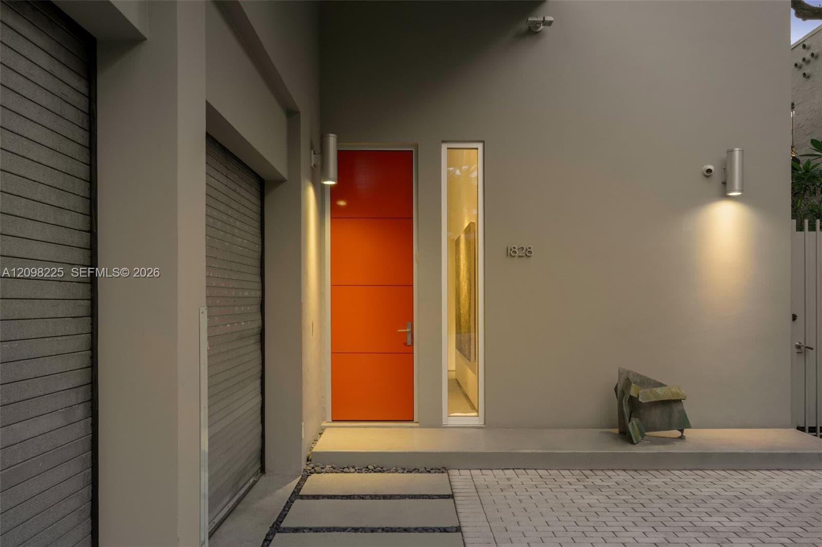 Modern architectural entry featuring a vibrant orange slab door, vertical transom window, dual cylinder wall-mounted exterior light fixtures, and a paved walkway with concrete pavers