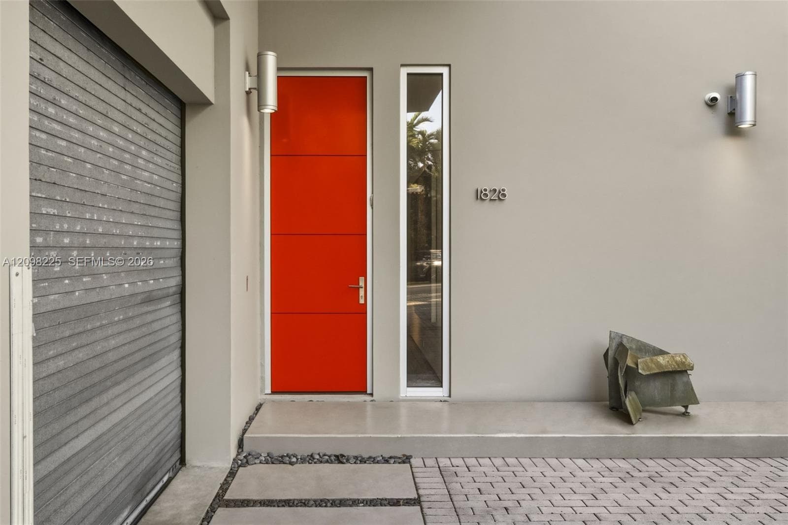 Modern exterior entry featuring a high-contrast orange door, tall side-light window, metallic wall-mounted light fixtures, and a paved driveway