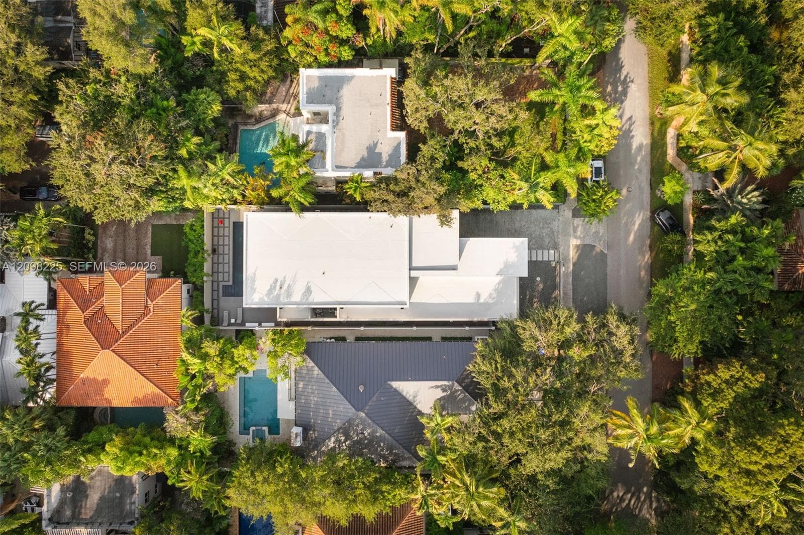 Aerial view of a property featuring a white flat-roofed residence, a swimming pool with an integrated spa, paved hardscaping, and mature perimeter landscaping