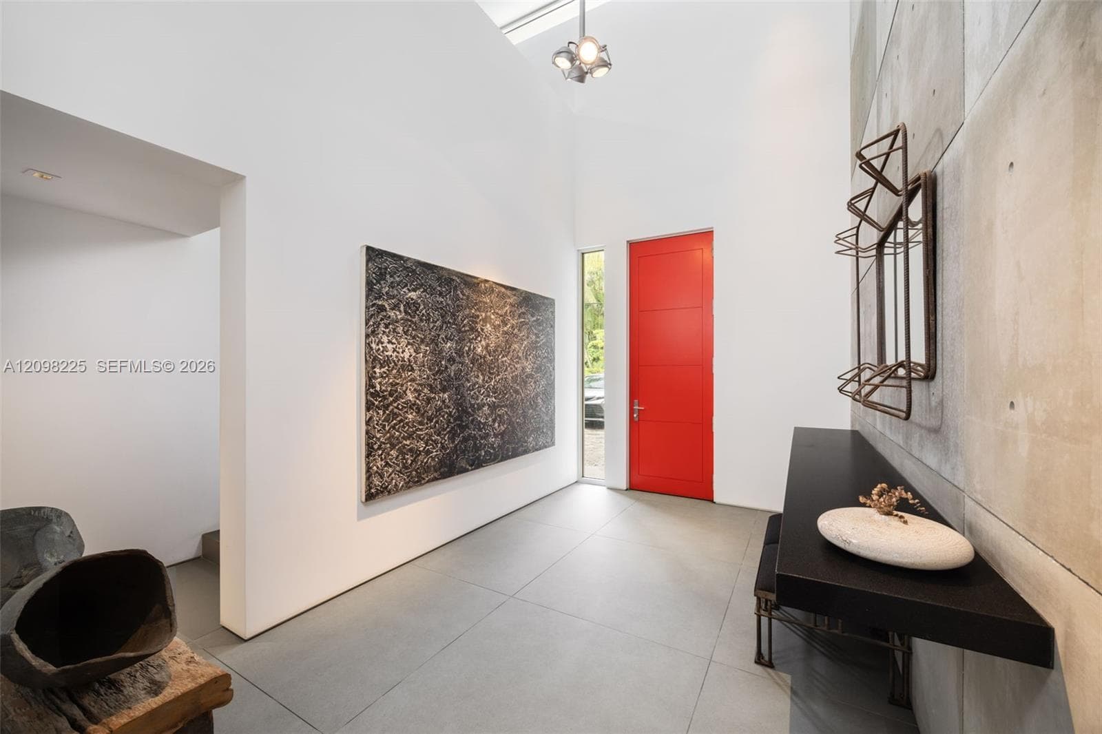 High-ceiling entryway featuring large-format tile flooring, a prominent red entry door, a floor-to-ceiling vertical window, and a feature wall composed of modular concrete panels with a built-in black console shelf