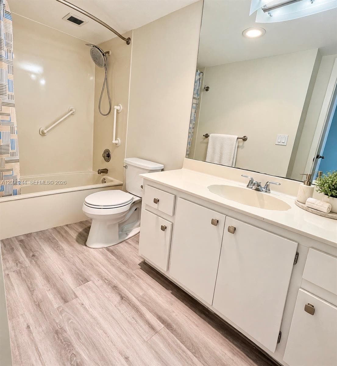 Bathroom featuring a large vanity with a solid-surface countertop, integrated basin, wood-finish flooring, and a shower-tub combination with wall-mounted grab bars and a handheld fixture