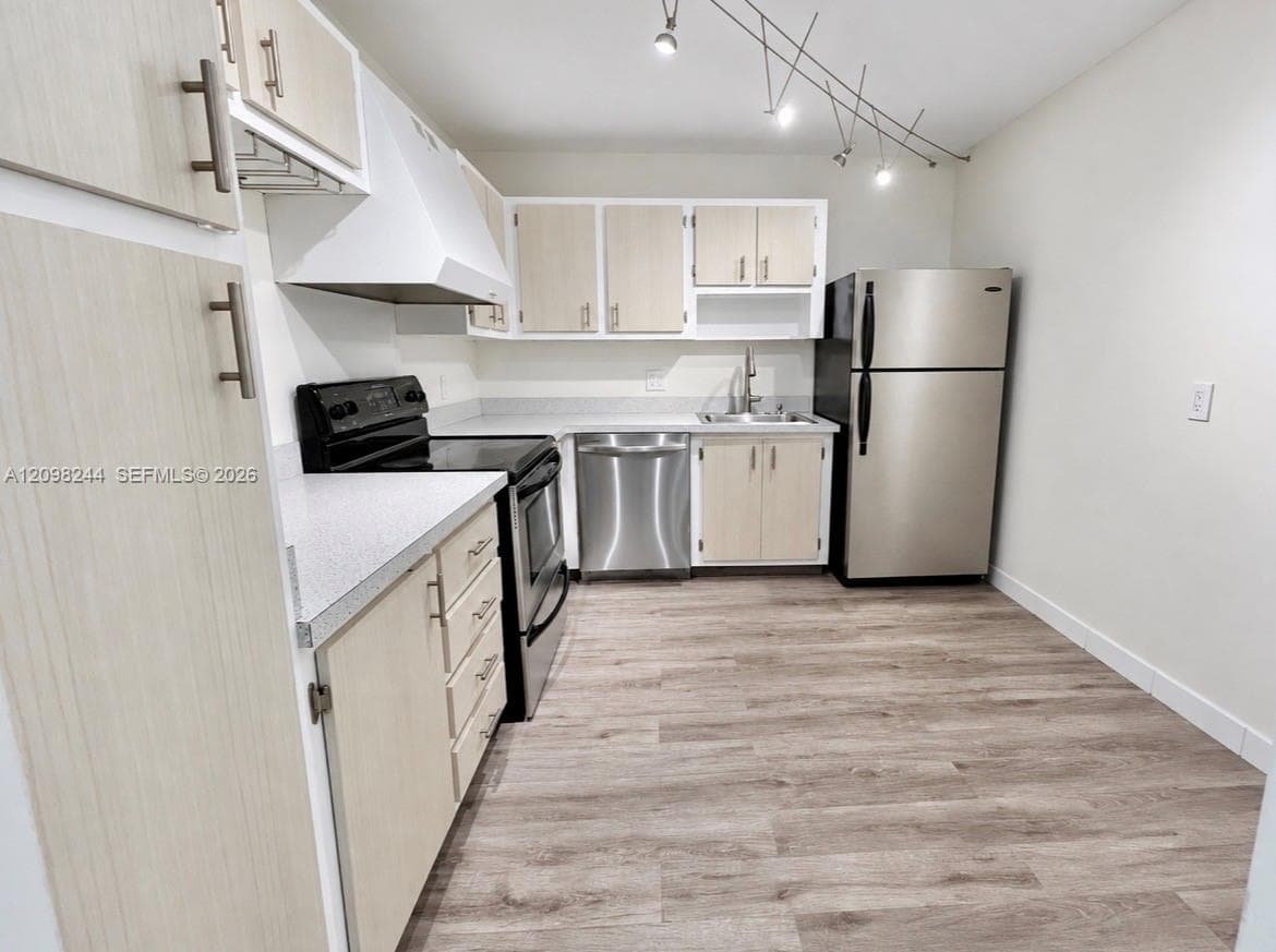Kitchen featuring light wood-finish cabinetry, stainless steel appliances, solid surface countertops, track lighting, and wood-finish flooring