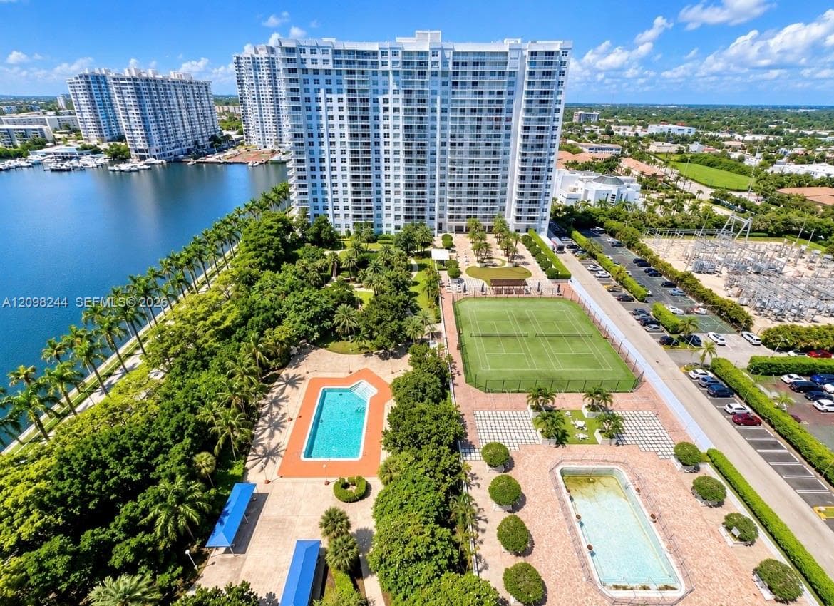 High-rise residential building featuring multiple swimming pools, a tennis court, landscaped grounds, and water frontage
