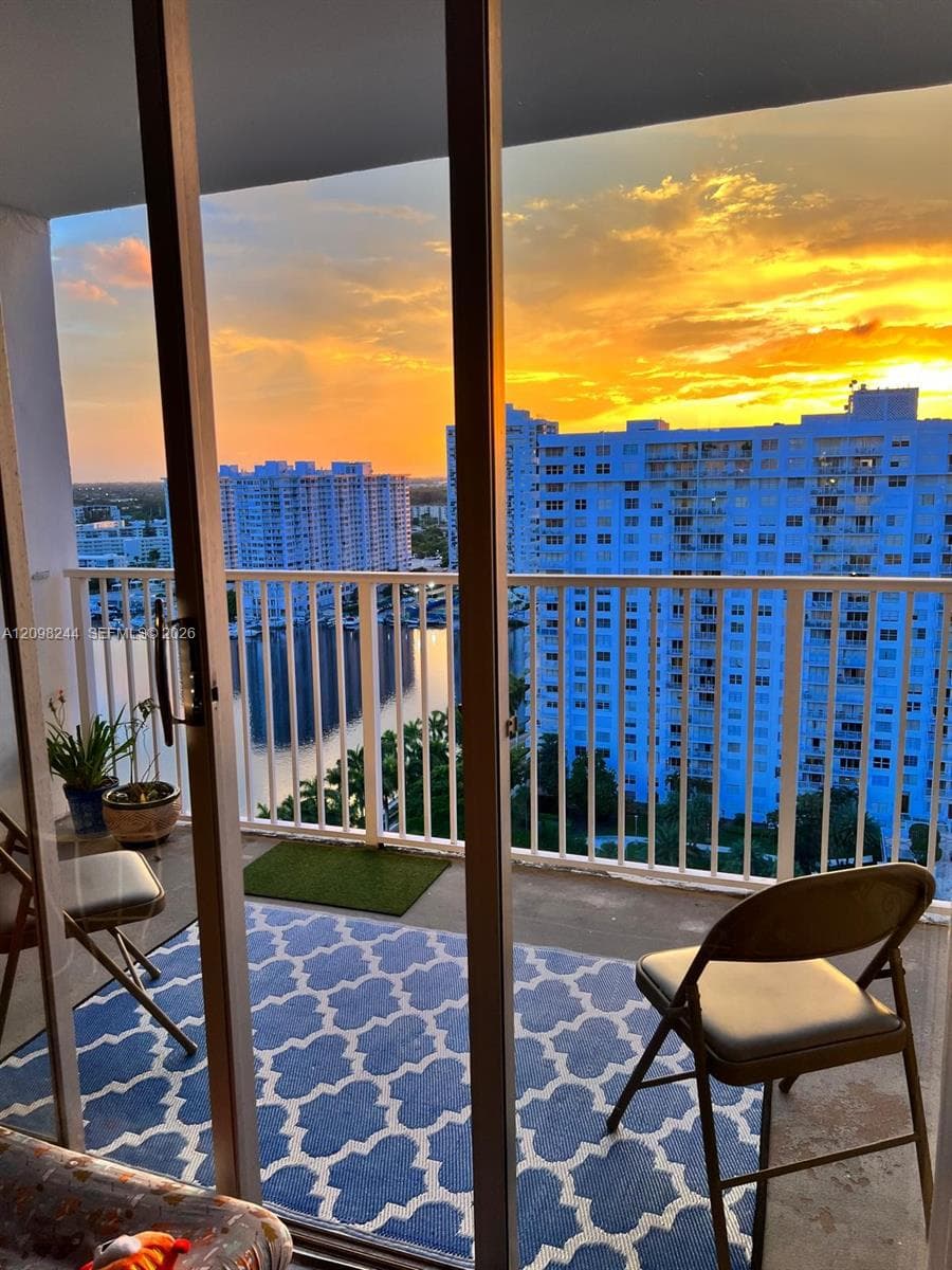 Private balcony featuring metal railings and concrete flooring with a backdrop of urban high-rises, waterfront scenery, and sunset skies