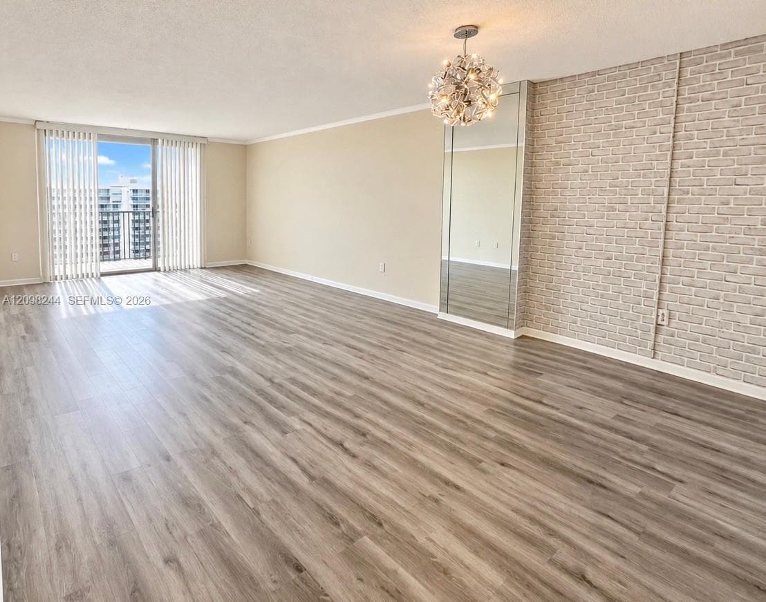 Living area featuring wood-finish flooring, a textured brick accent wall, a floor-to-ceiling mirror panel, a decorative chandelier, and sliding glass balcony doors