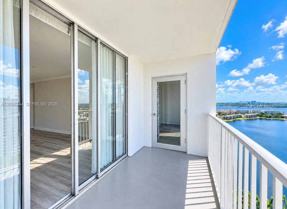 Covered balcony featuring a white metal railing, solid surface flooring, and a glass-paned door, complemented by large sliding glass doors and expansive water scenery