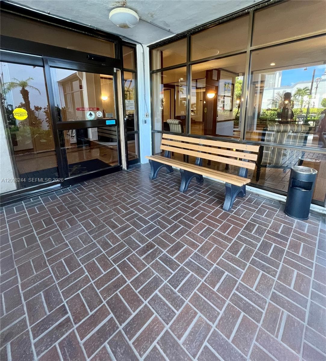 Covered building entrance featuring a herringbone-patterned tile floor, expansive floor-to-ceiling glass wall, and aluminum-framed sliding glass doors