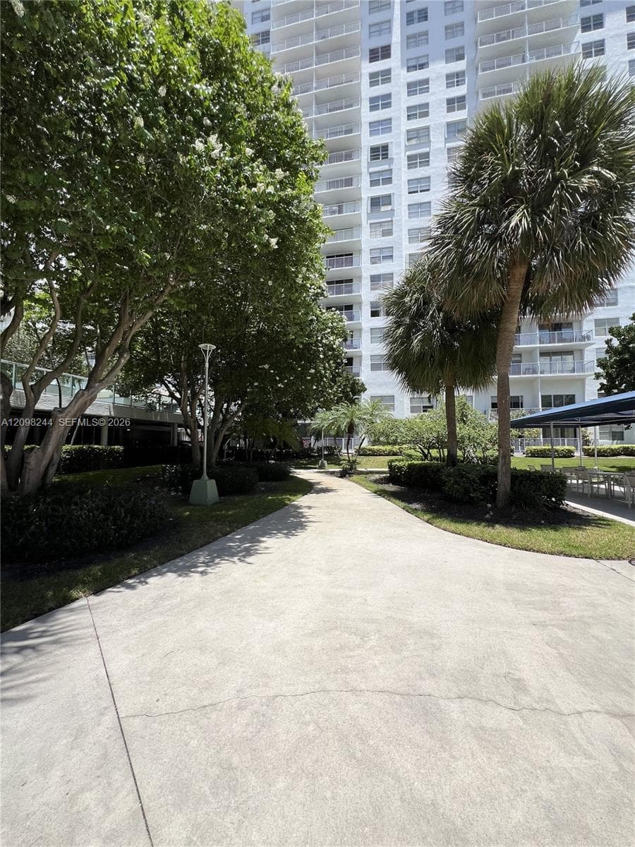 Paved walkway surrounded by mature trees, palm trees, manicured landscaping, and exterior pole lighting