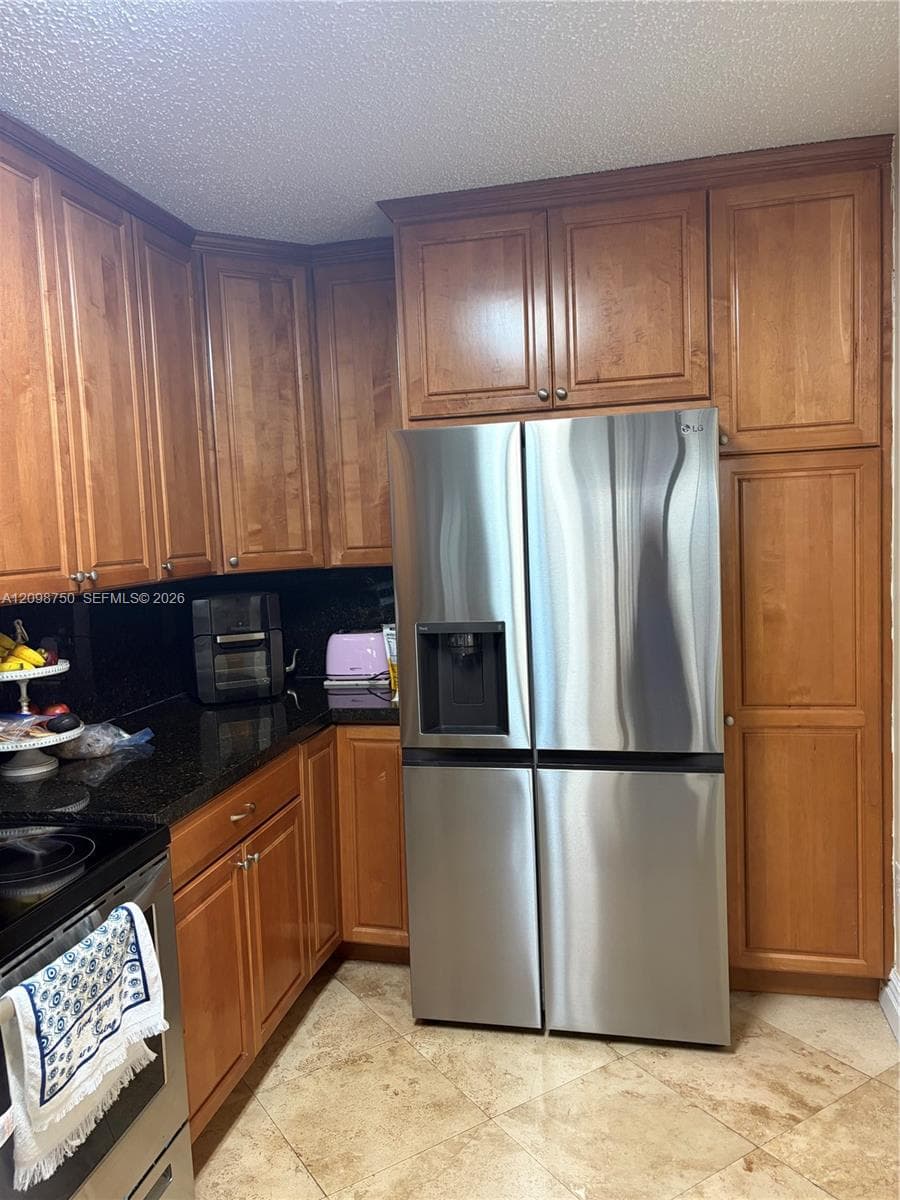 Kitchen featuring medium-tone wood cabinetry, dark stone countertops, stone-finish tile flooring, and a stainless steel four-door refrigerator
