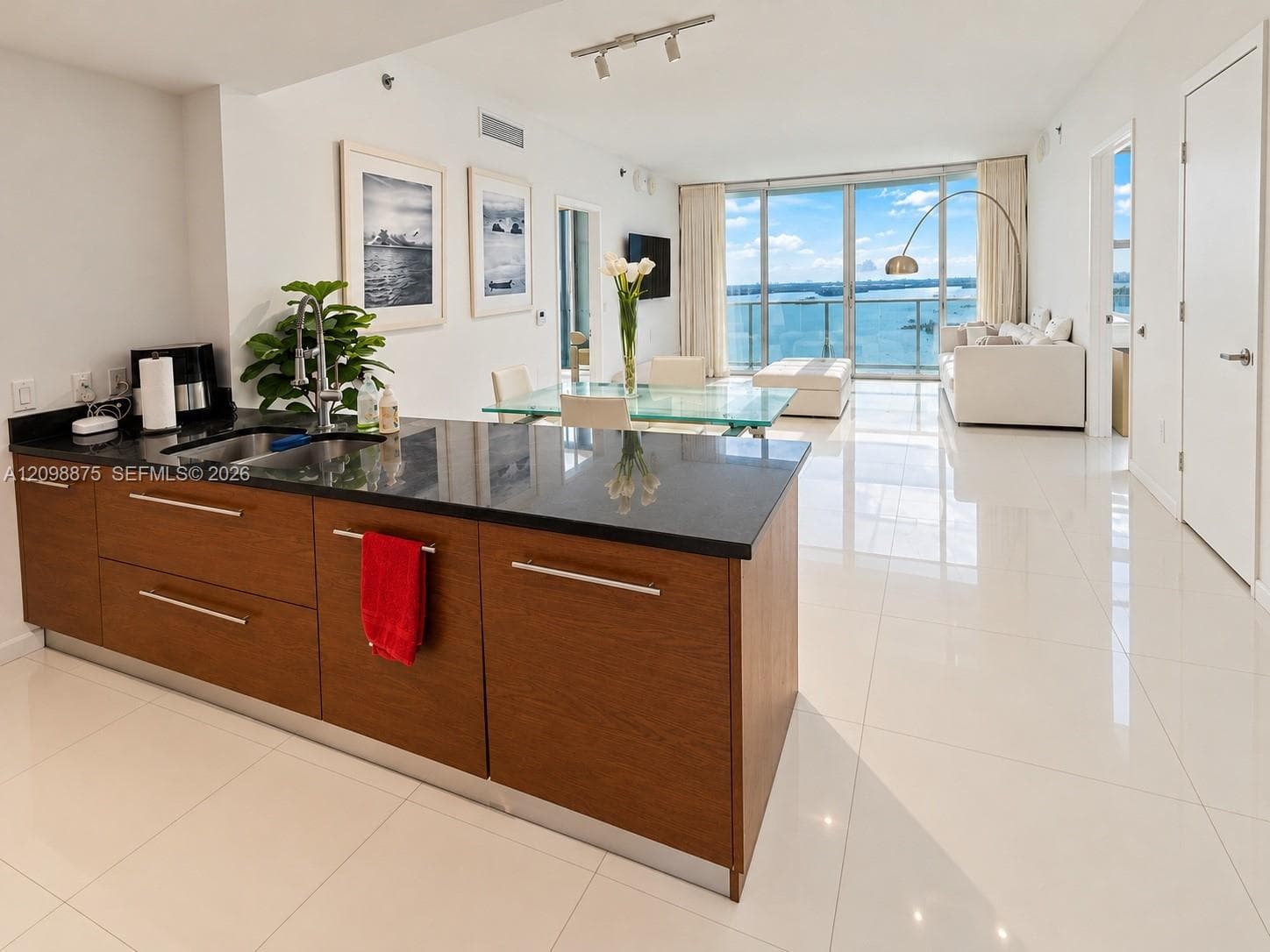 Open-concept living space with expansive floor-to-ceiling windows, waterfront scenery, sleek kitchen island featuring a dark countertop, and polished tile flooring