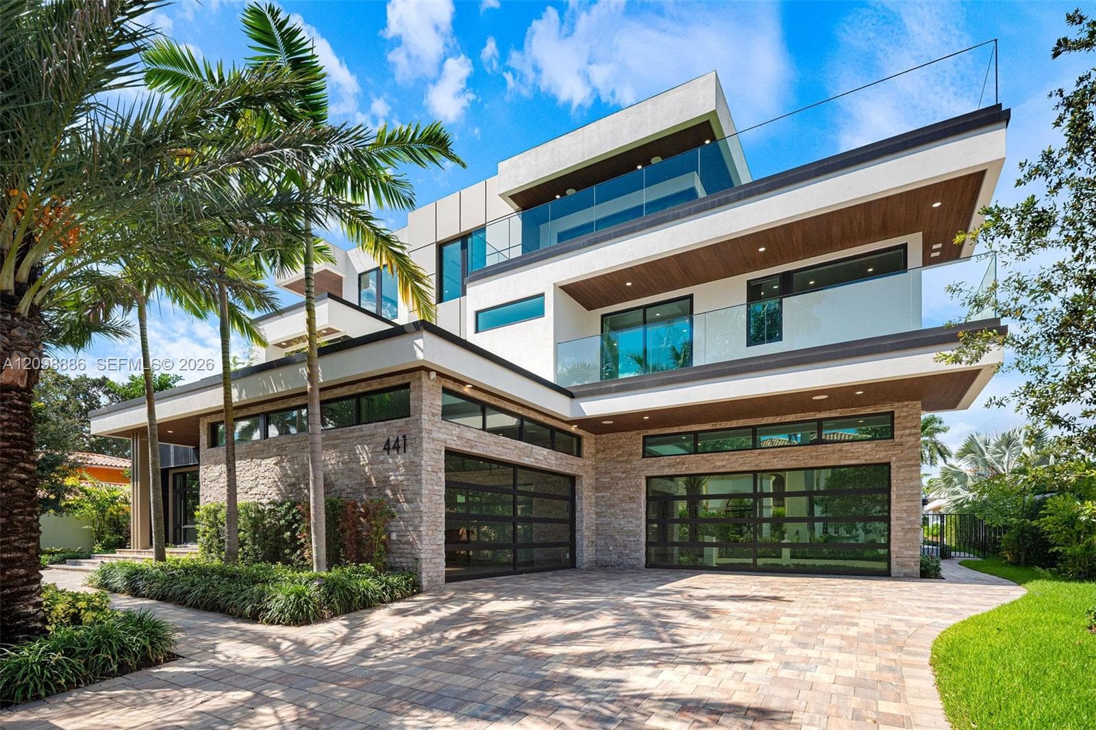 Modern multi-level exterior featuring stacked stone masonry, horizontal glass panel garage doors, glass balcony railings, and wood-finish soffits